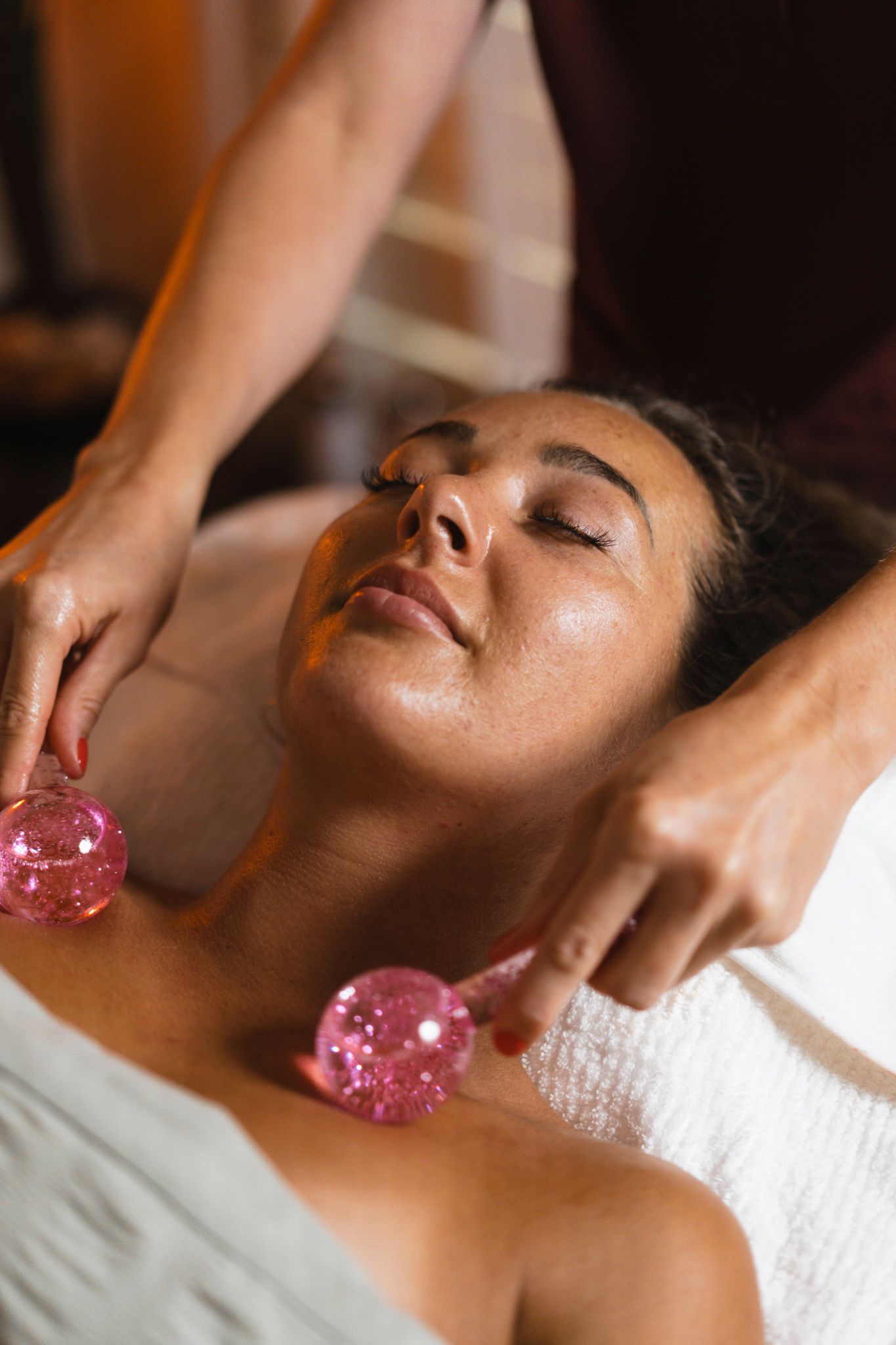 A relaxed spa client receives a facial massage treatment with pink crystal massage tools on white towels.