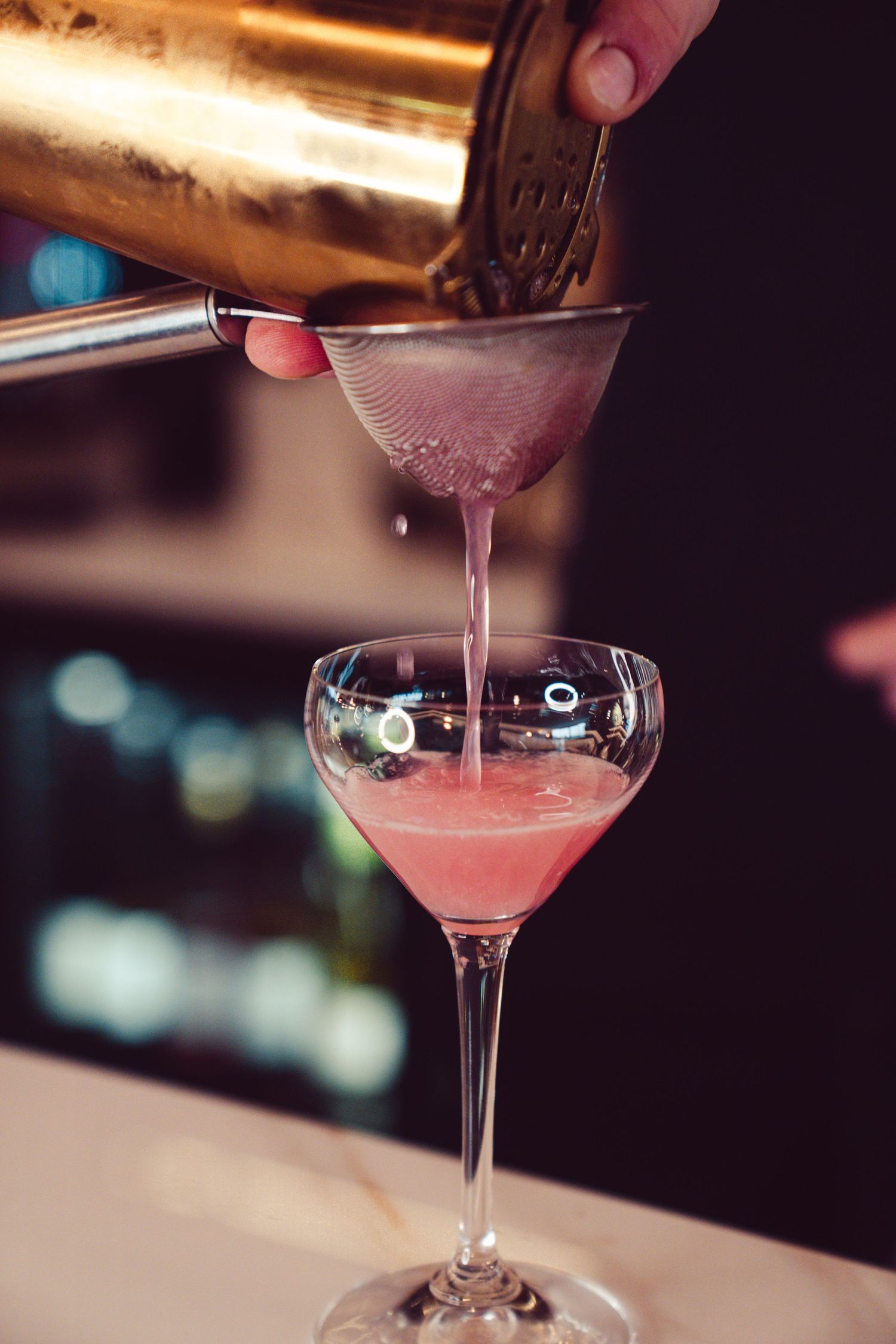 Pink cocktail being poured into a martini glass at an upscale bar with bokeh lighting in the background.