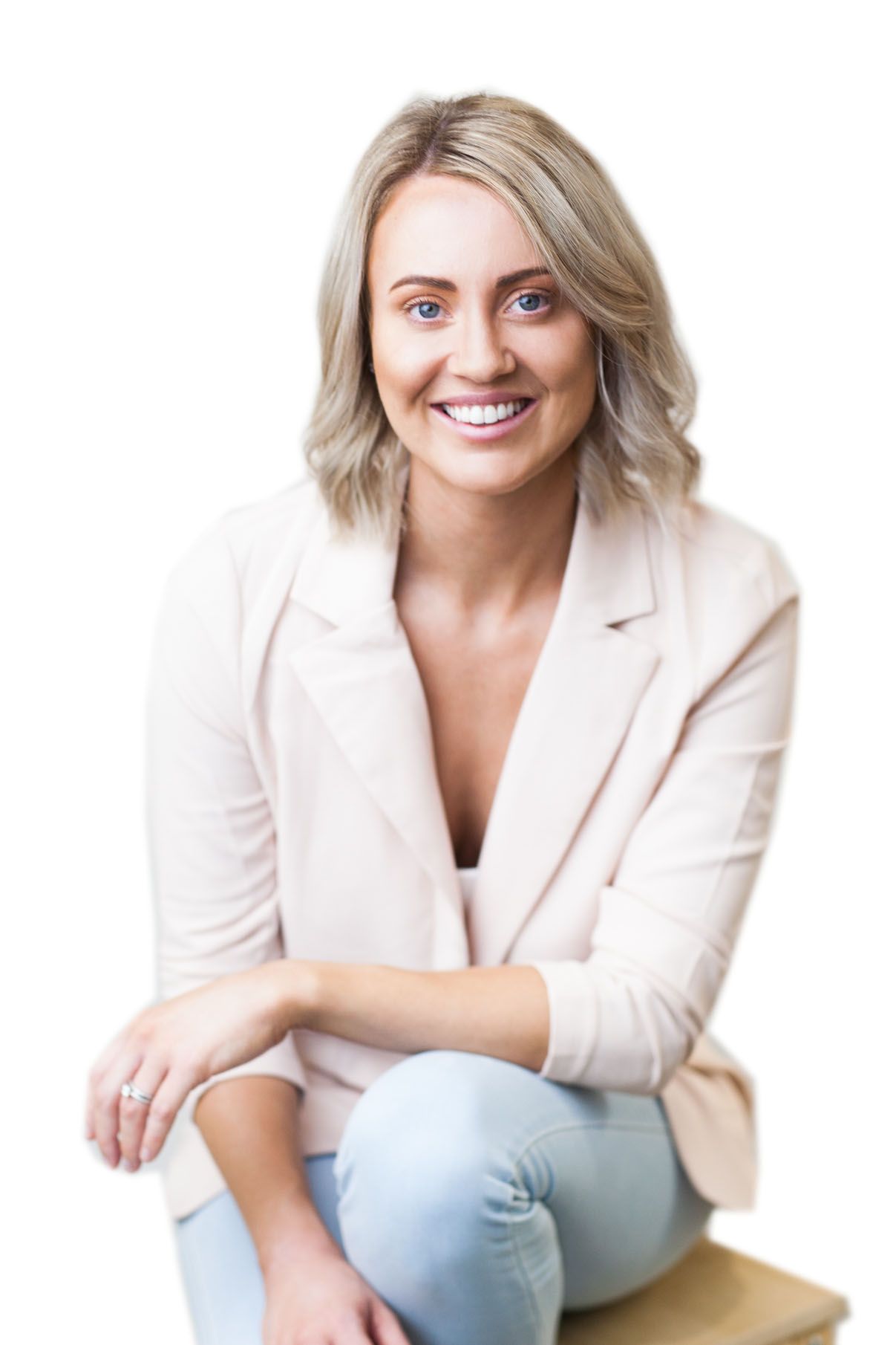 Smiling blonde woman in light pink blazer and jeans sitting casually against a white background.