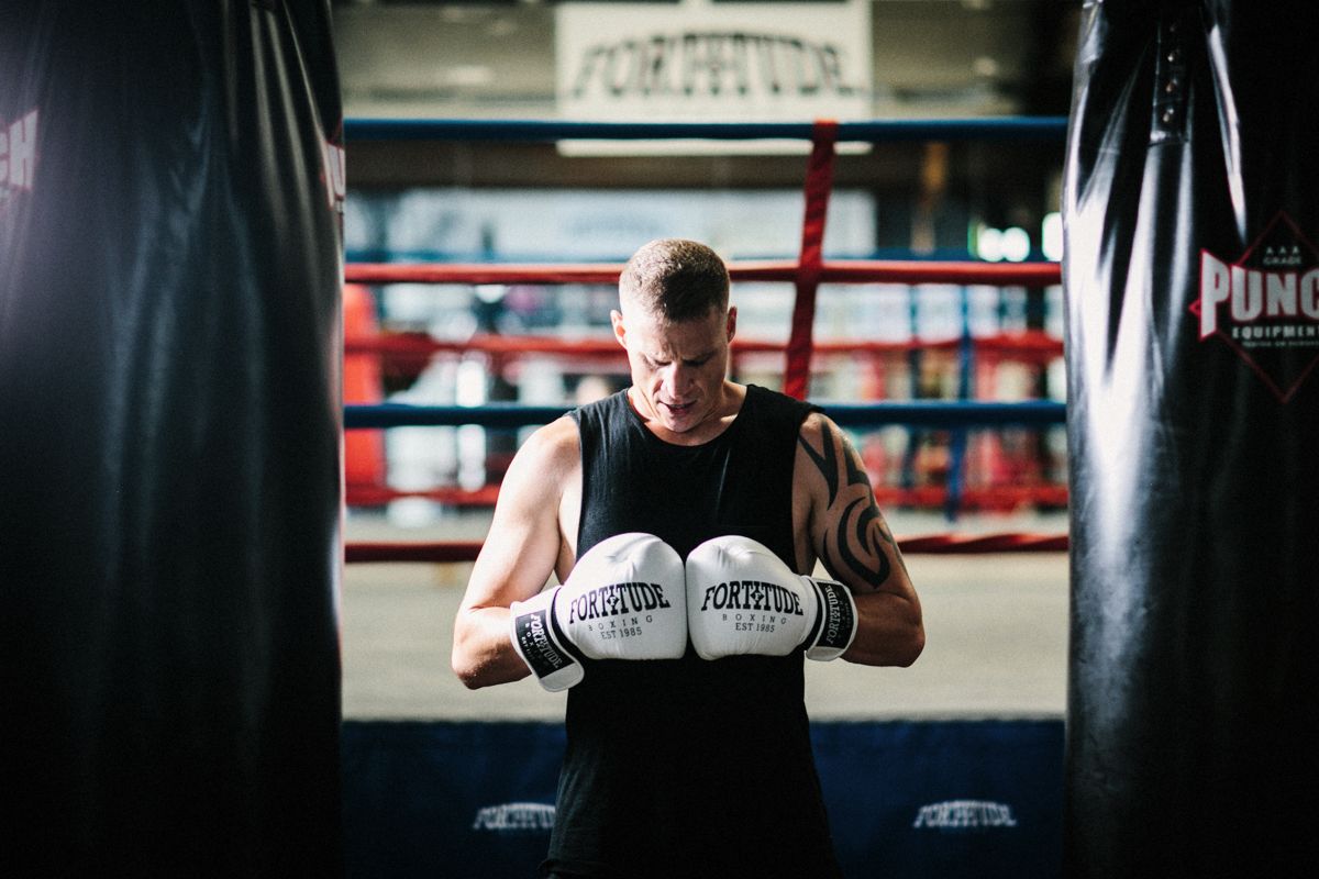 Boxer in black tank top wearing white boxing gloves trains in a gym near a heavy bag and boxing ring.