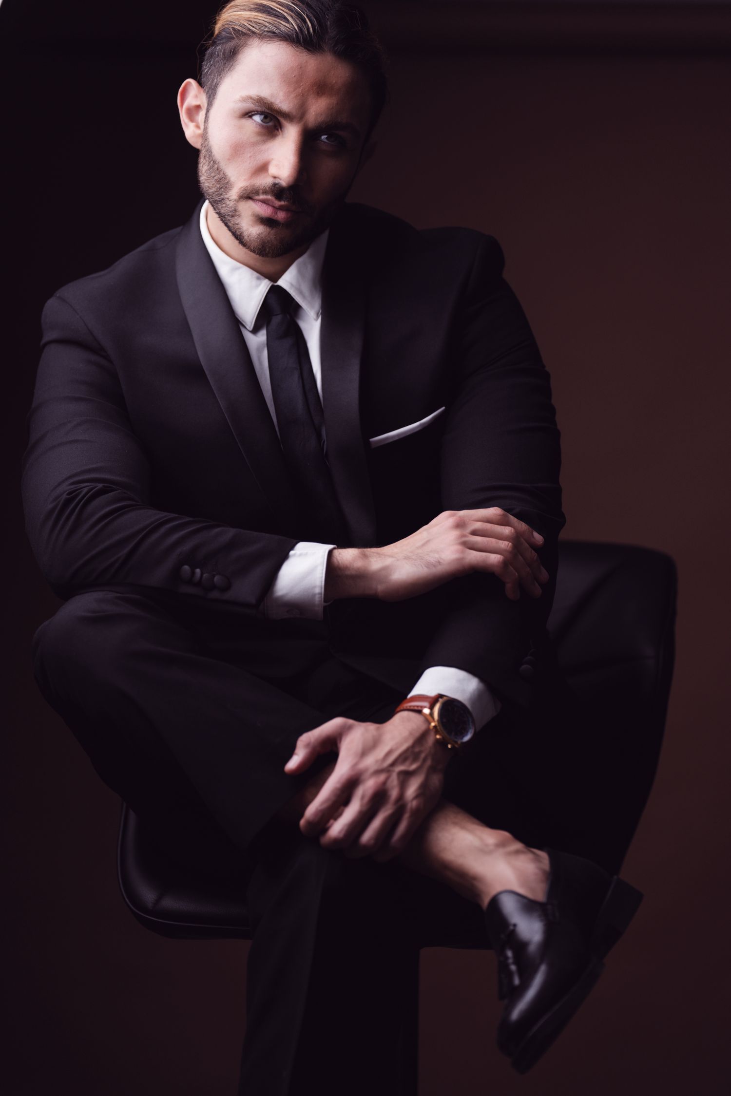 Elegant man in black suit and tie sitting confidently, wearing a gold watch, against a dark background.