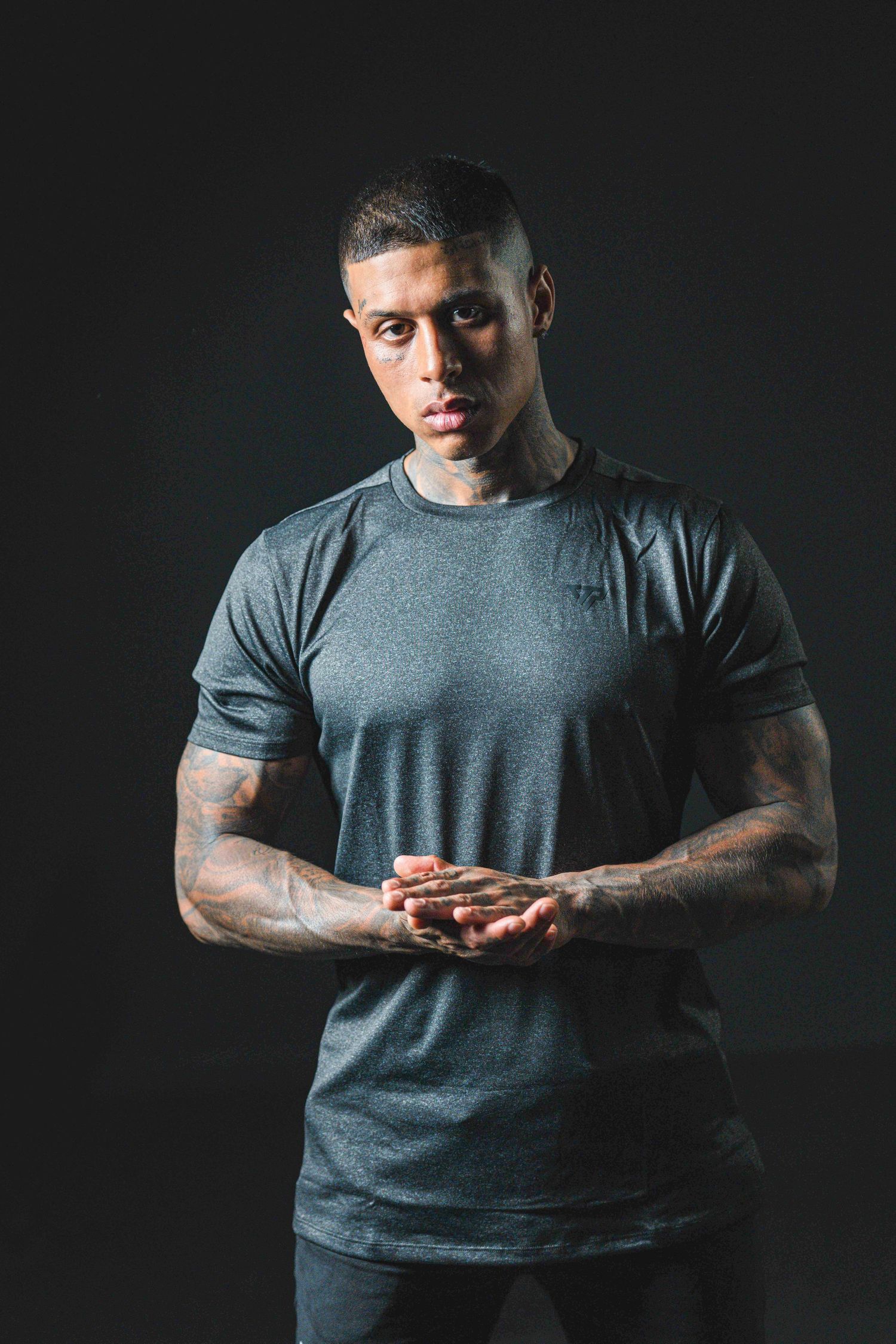 Tattooed muscular man in gray t-shirt poses against dark background with hands clasped together.