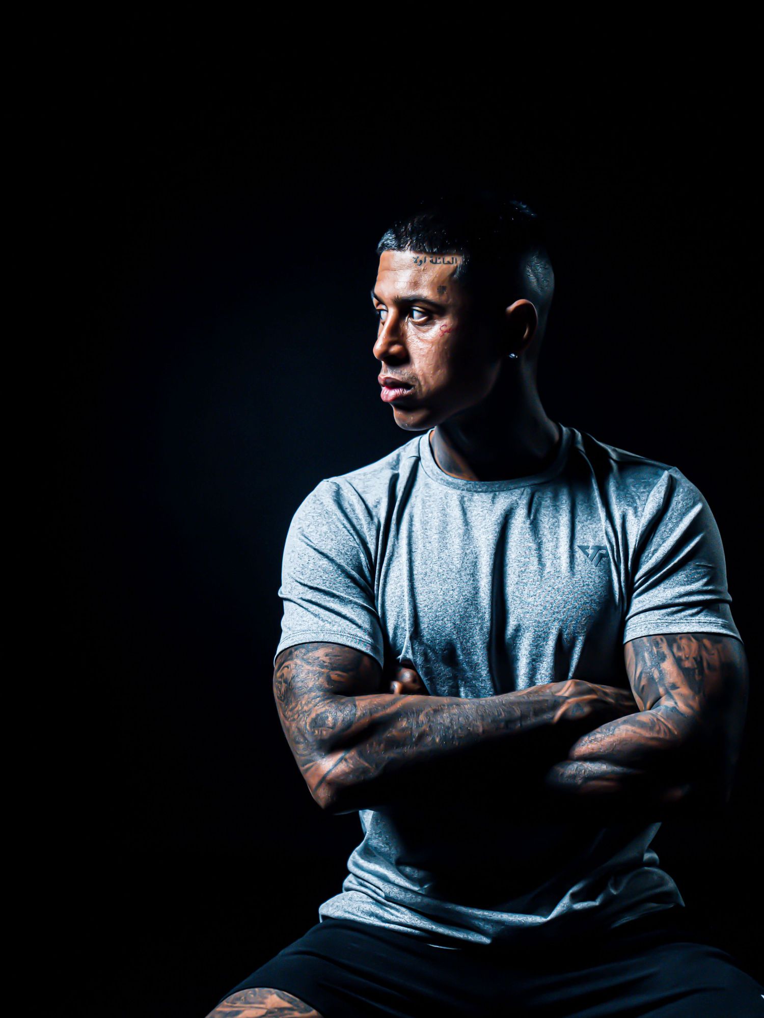 Muscular man with tattoos sits cross-armed in gray t-shirt against dark background in dramatic studio lighting.