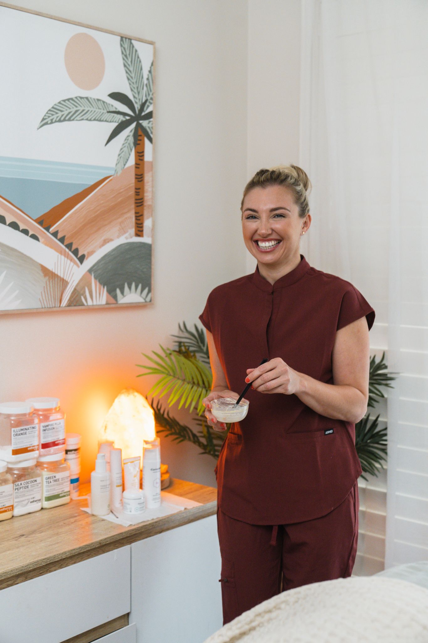 A person in burgundy scrubs stands in a medical office with palm tree artwork and product displays.
