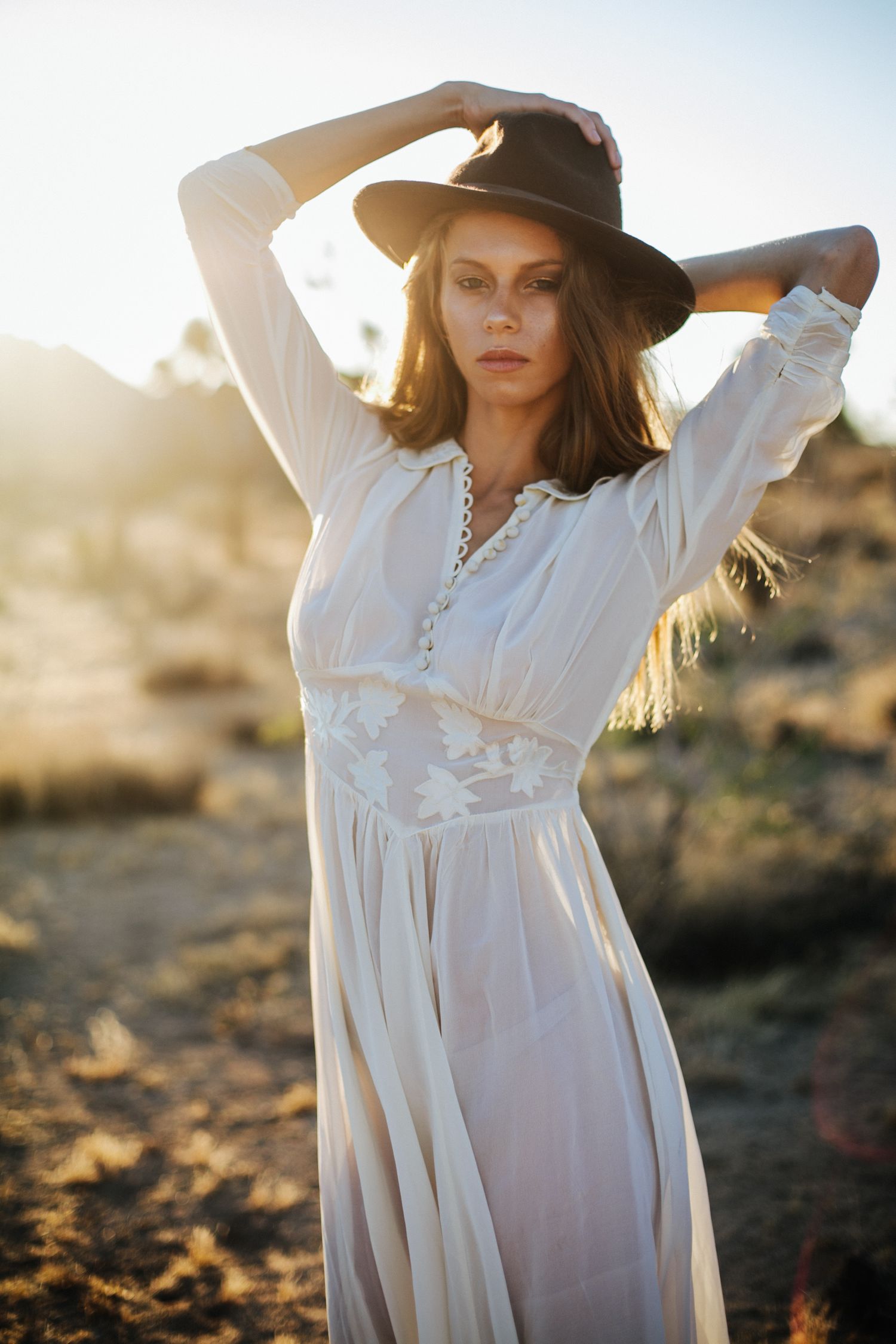 Ethereal desert photoshoot featuring flowing white dress and wide-brimmed hat at sunset.