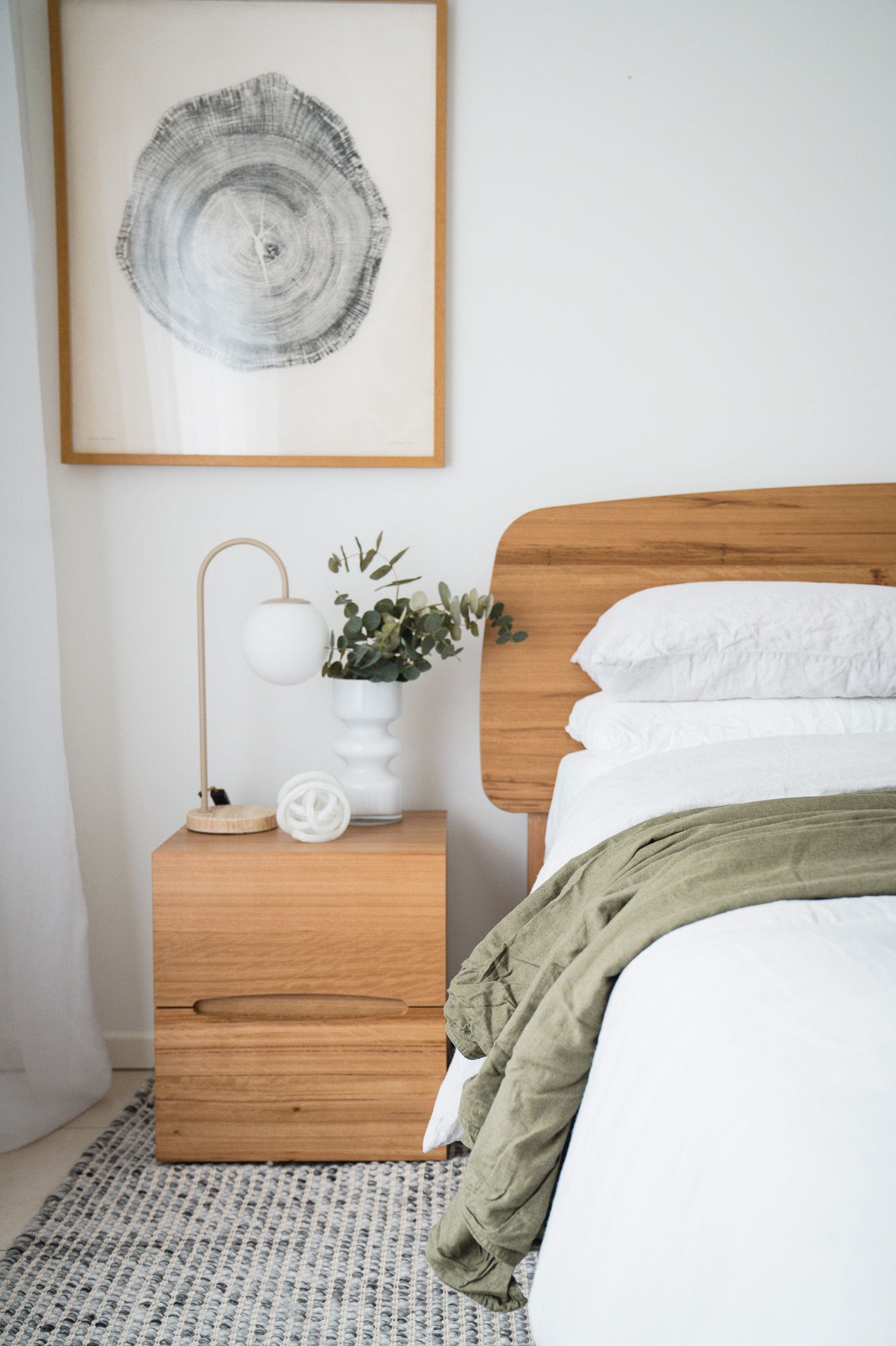 Minimalist bedroom with wooden headboard, nightstand, white bedding, olive green throw, eucalyptus stems, and tree ring wall art.