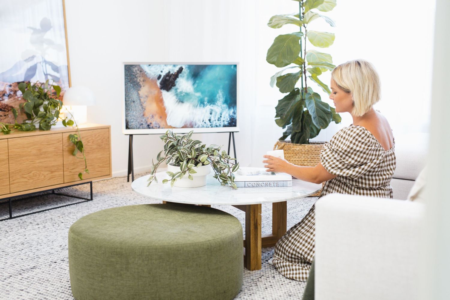 Woman relaxing in modern living room with Samsung The Frame TV displaying ocean art, green ottoman, and tropical plants.