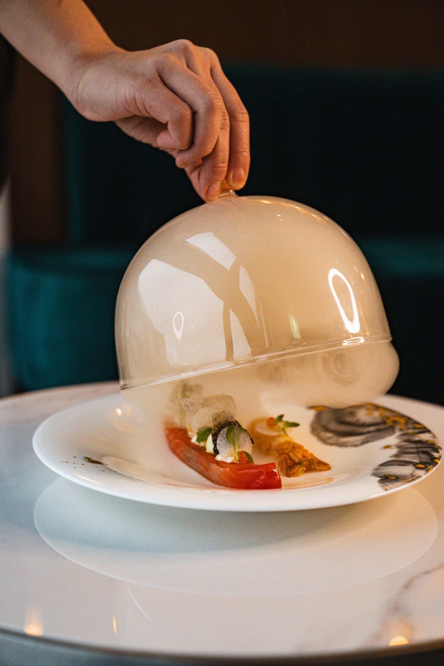 A hand lifts a white cloche dome to reveal a plated dish with colorful ingredients on an elegant white table setting.