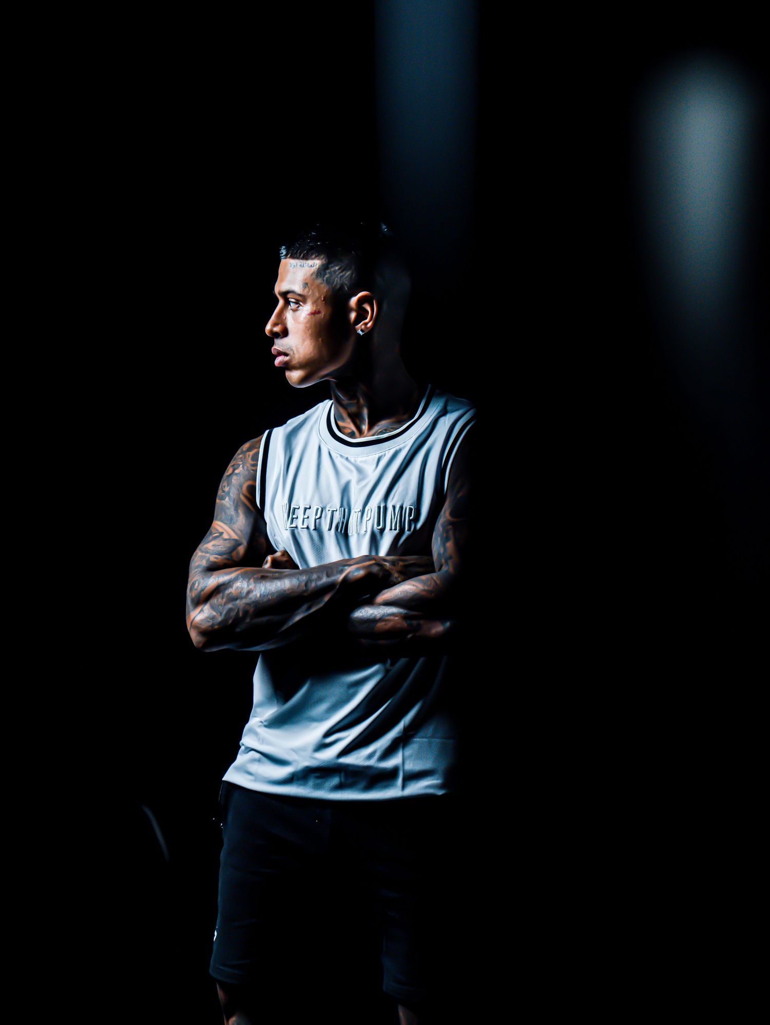 Tattooed man in white sleeveless shirt stands arms crossed in dramatic dark studio lighting.