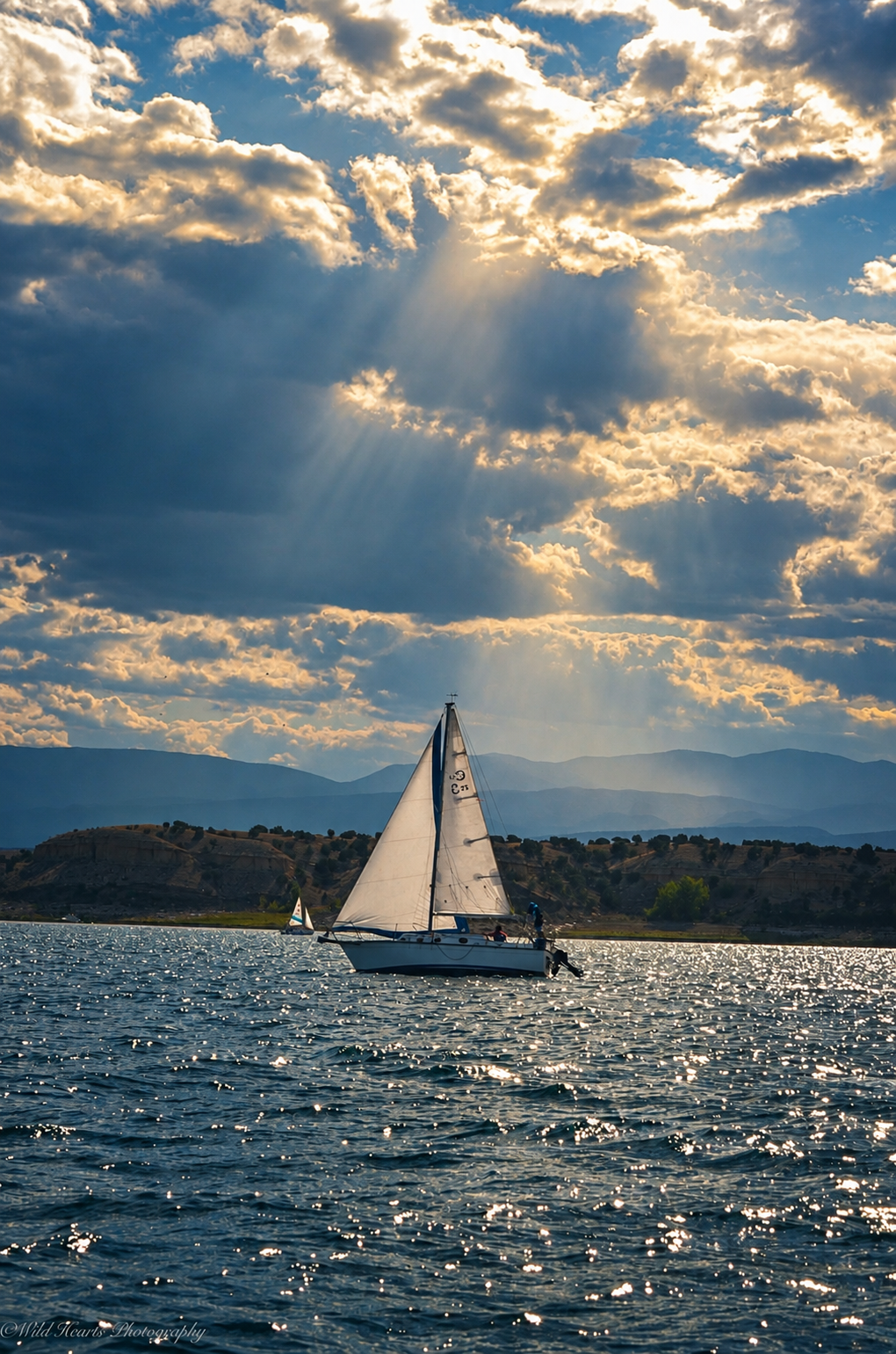 Peaceful sailboat crossing a lake beneath dramatic clouds as golden sunlight breaks through the sky and reflects on the water.