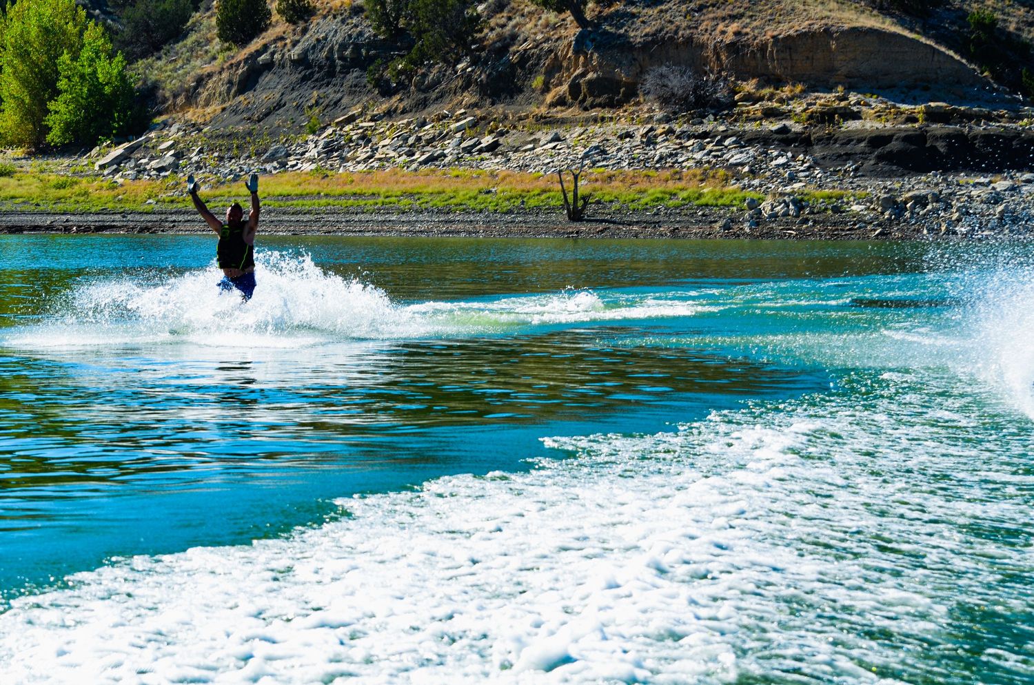 Wide-angle action photograph of a waterskier crossing a turquoise lake, leaving a trail of white spray with rocky hills behind.