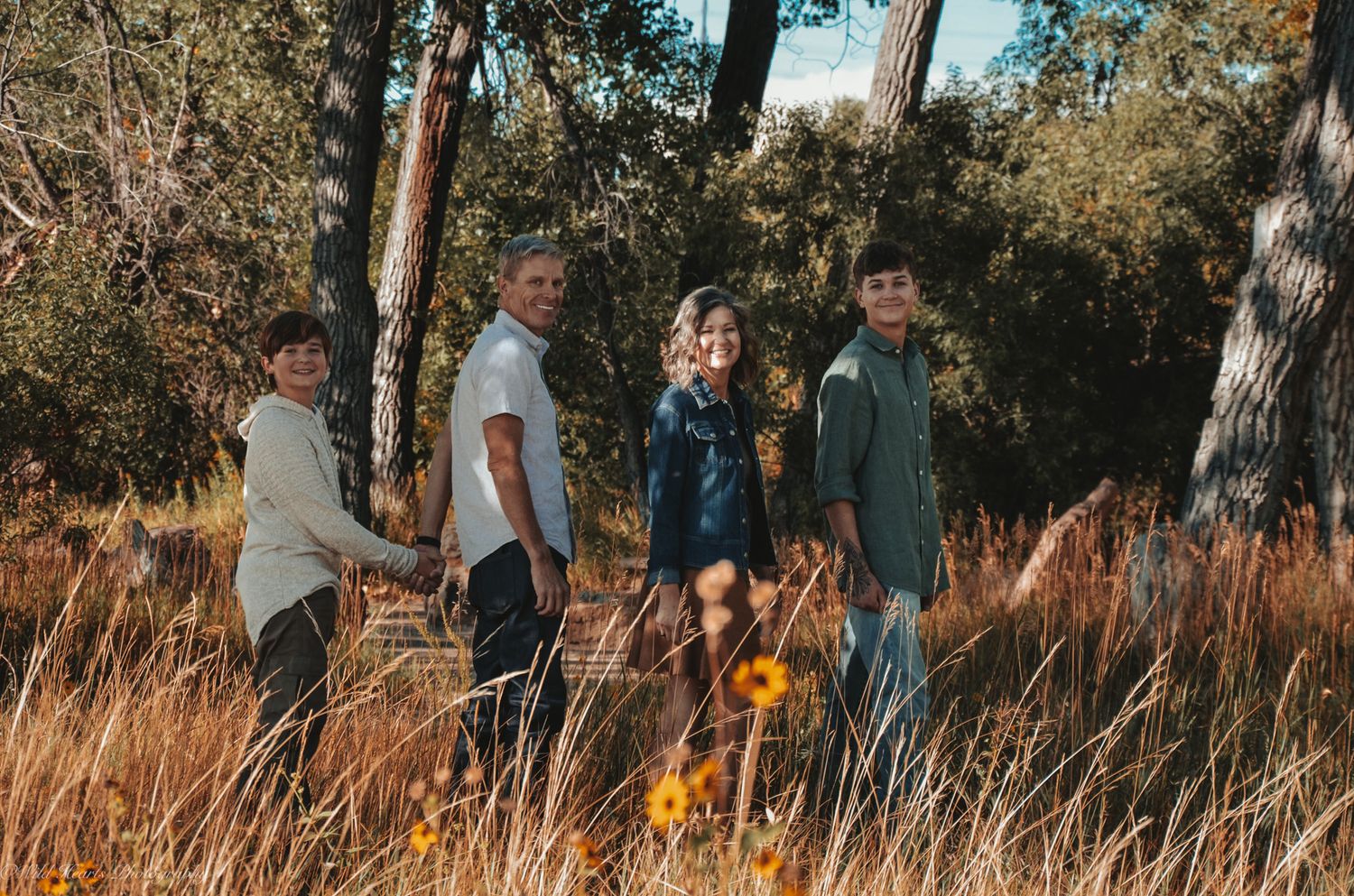 Outdoor fall family portrait of four standing together in tall golden grass surrounded by trees and warm autumn colors.