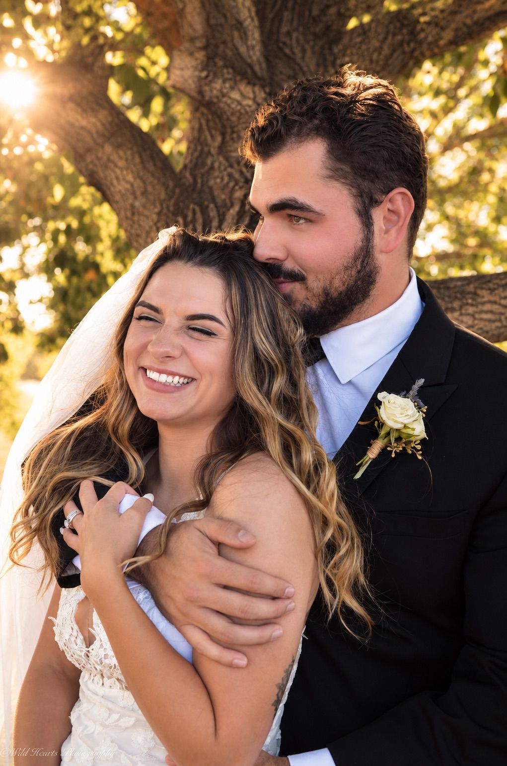 Romantic wedding portrait of a groom embracing his smiling bride from behind beneath golden autumn trees.