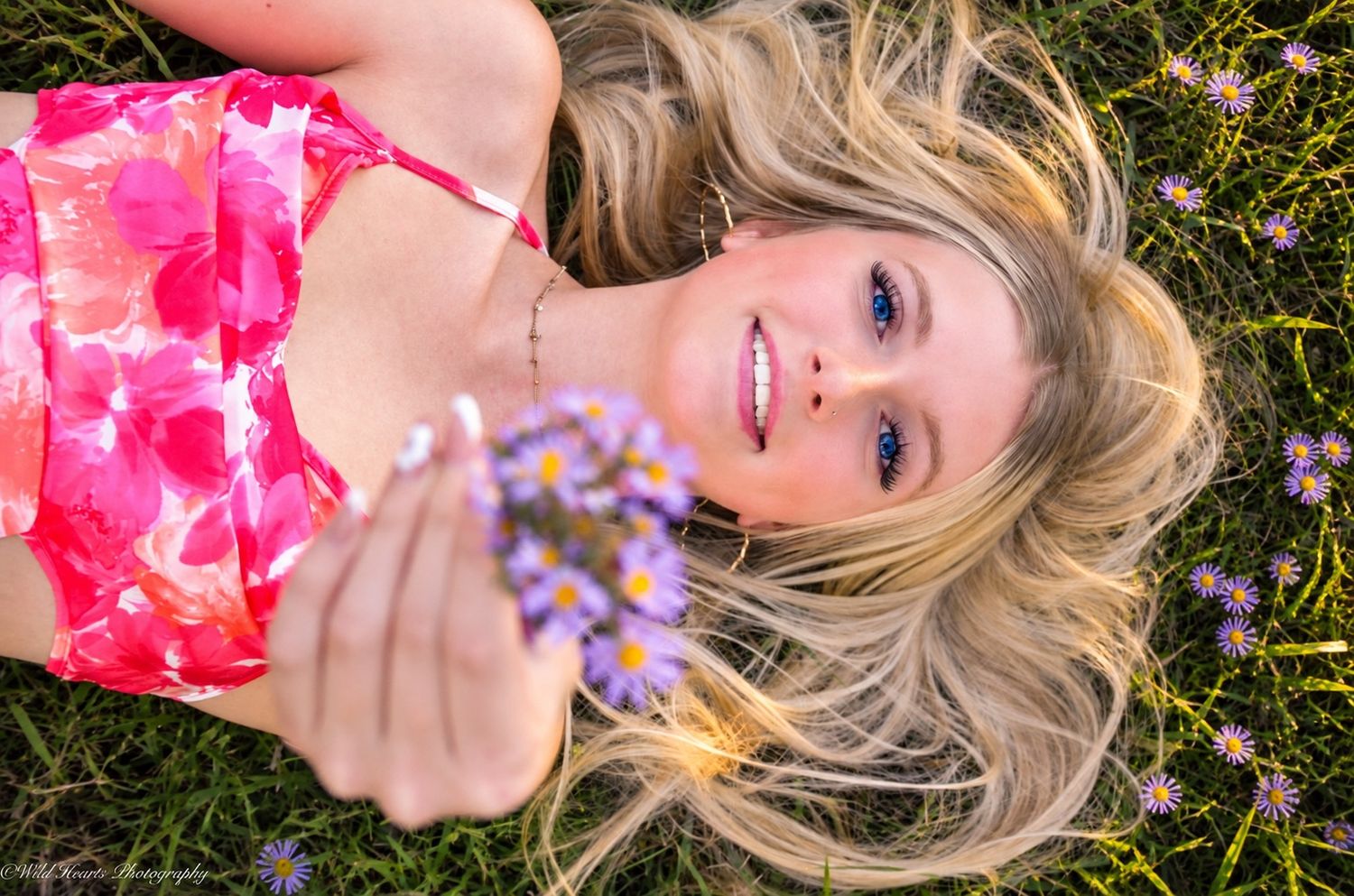 High school senior lying among purple wildflowers in a pink floral dress, holding flowers during a creative outdoor portrait session.