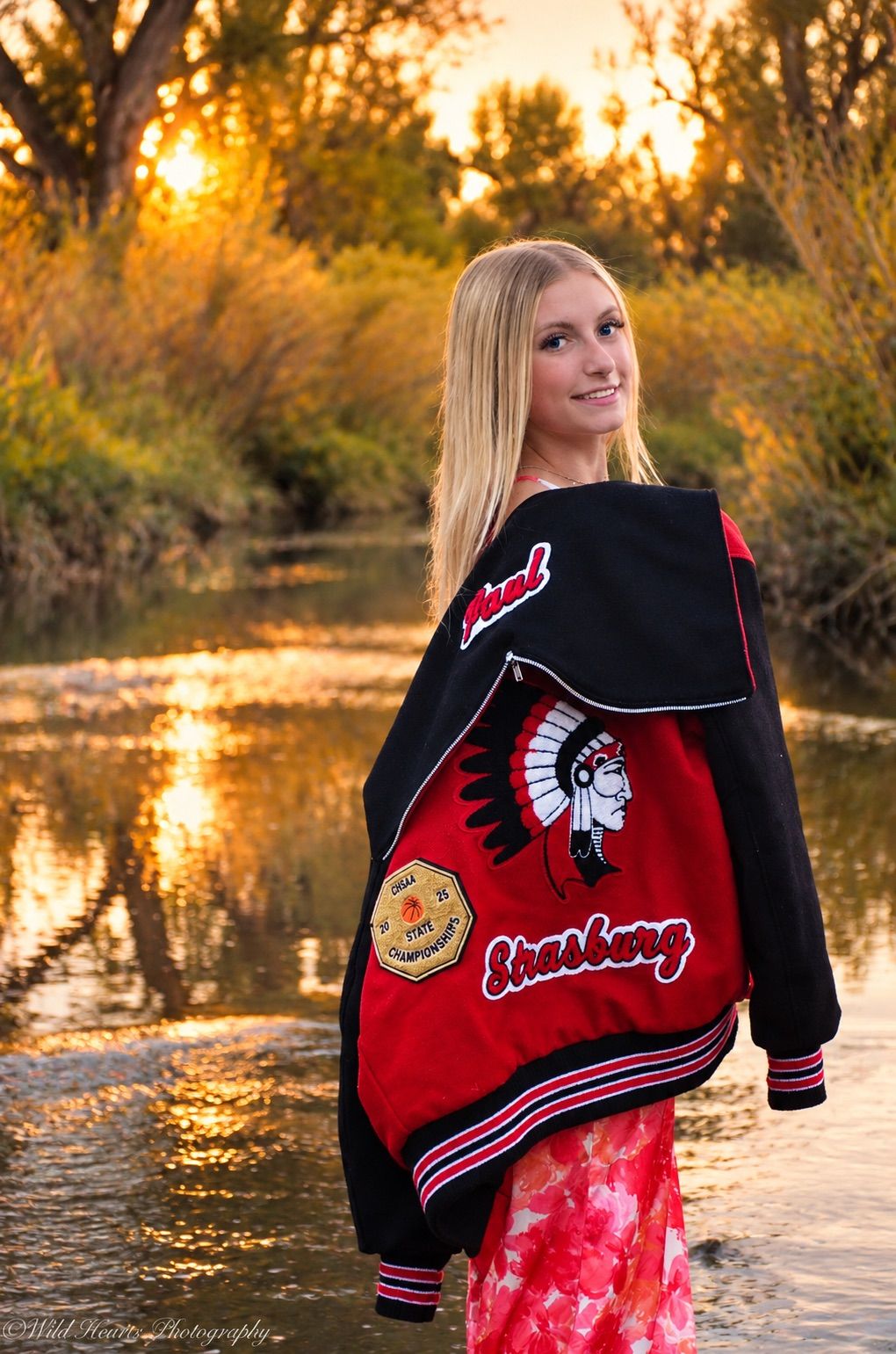 High school senior posing in a graduation stole over a red floral dress while standing in a creek during a golden-hour photo session.
