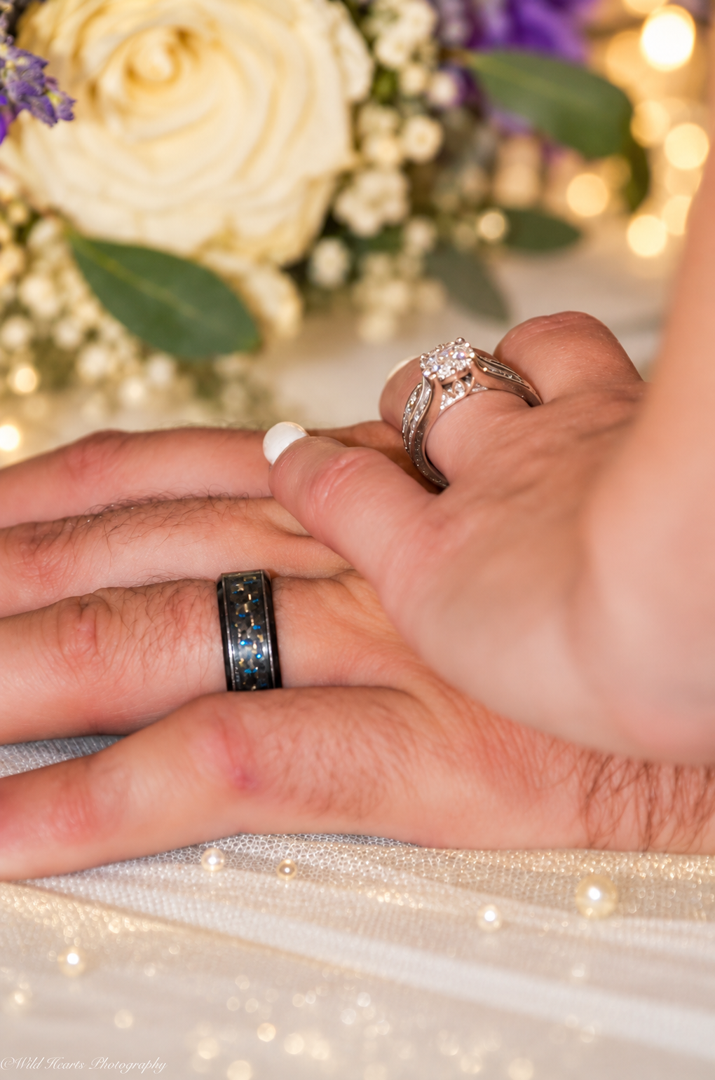 Close-up wedding photograph of a bride and groom's hands displaying their wedding rings beside a bouquet of white flowers.