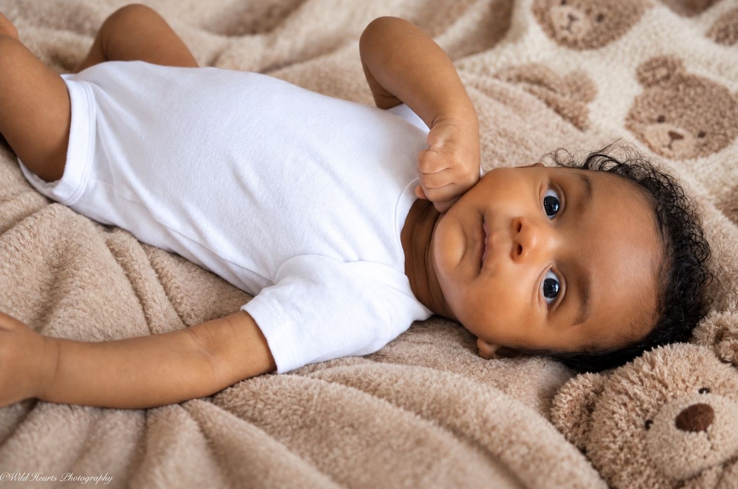 Sweet baby portrait of an infant wearing a white onesie while lying on a soft beige blanket and looking toward the camera.