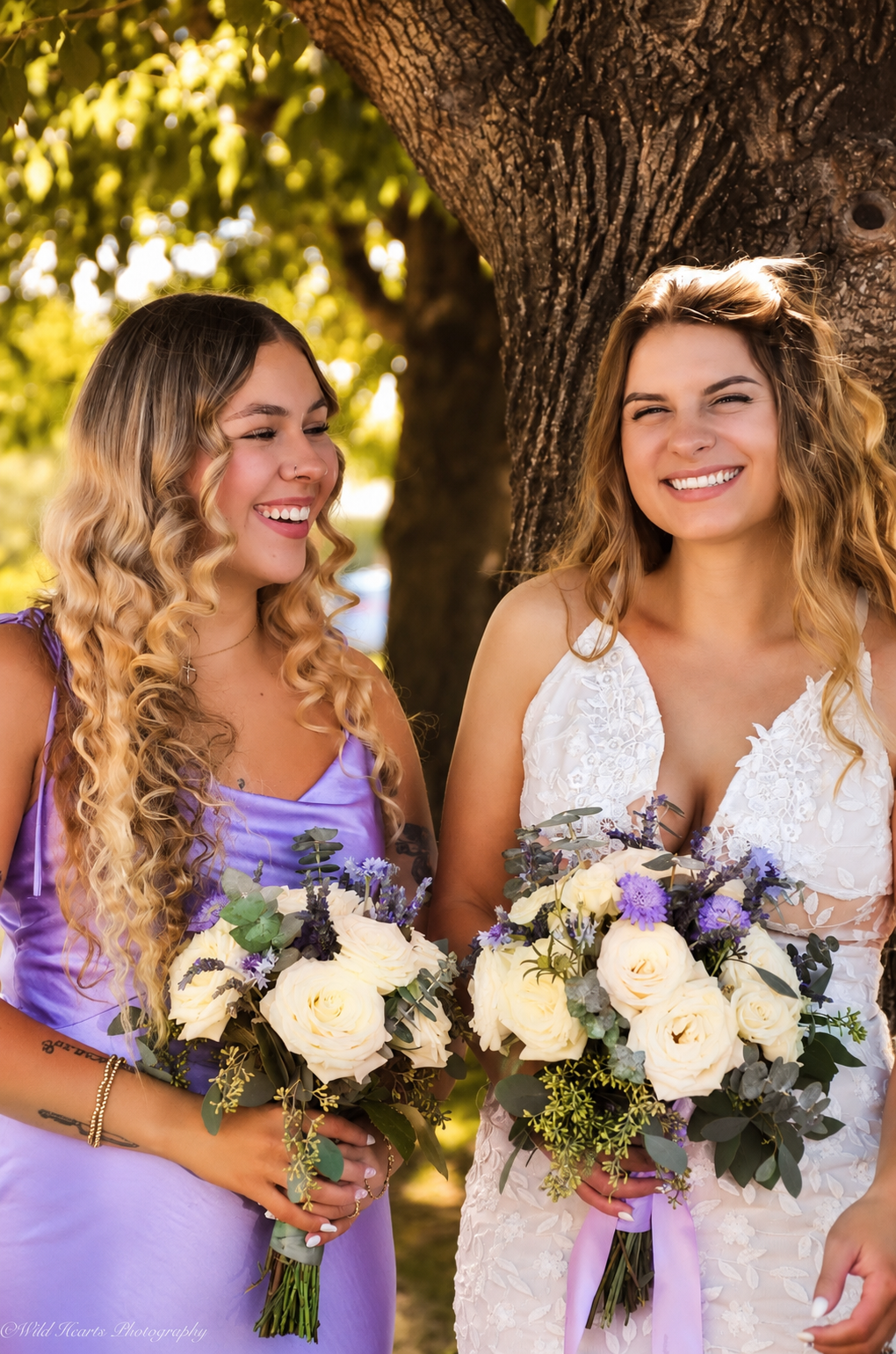 Bride in a white wedding gown standing beside her bridesmaid in a lavender dress, both holding floral bouquets beneath autumn trees.