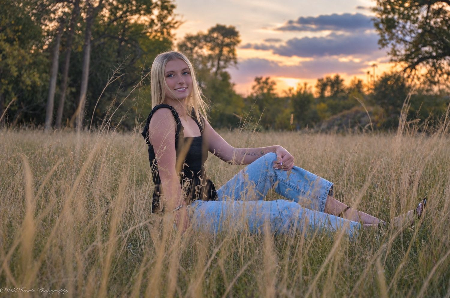 High school senior sitting in tall golden meadow grass wearing jeans and a black top during a beautiful sunset portrait session.