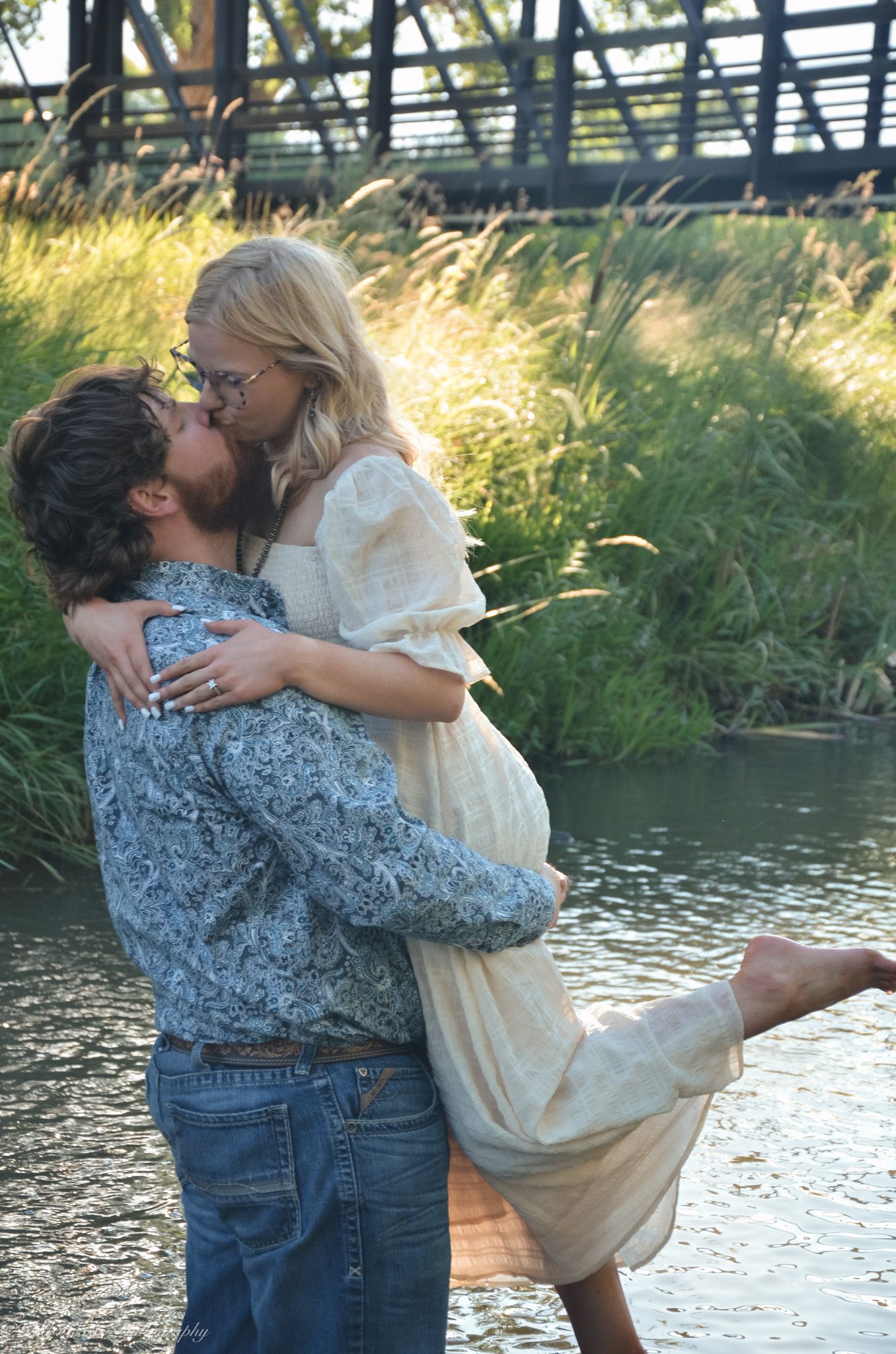 Romantic outdoor couples portrait of a man lifting and embracing his partner beside a creek surrounded by lush greenery.
