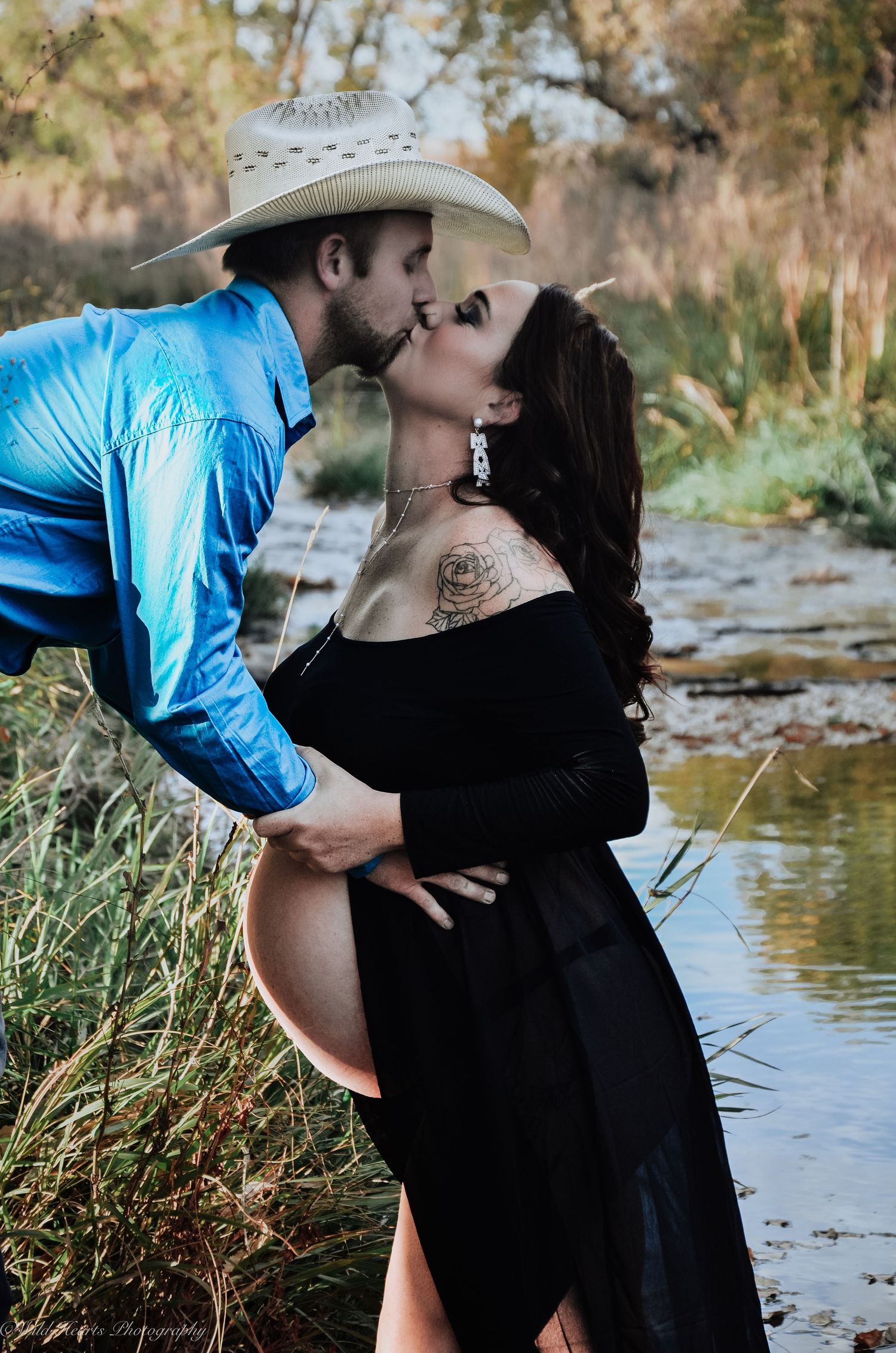 Expectant couple embracing beside a creek during an outdoor maternity photography session, with the mother wearing a black dress.