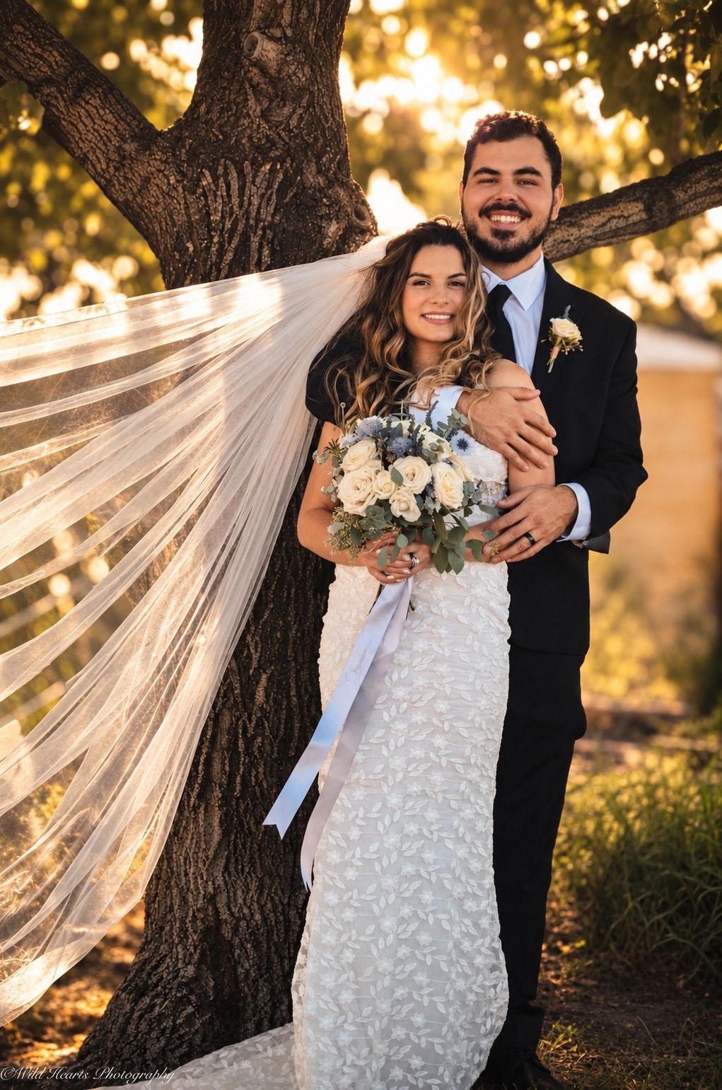 Bride in a white wedding gown holding a bouquet while standing with her groom beneath autumn trees during a romantic outdoor wedding.