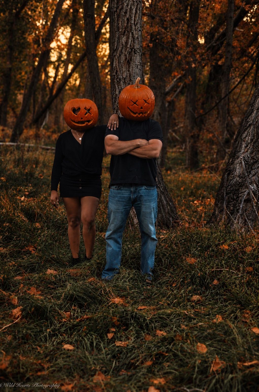 Dark romantic Halloween couples portrait featuring two people wearing carved pumpkin heads in a moody autumn forest.