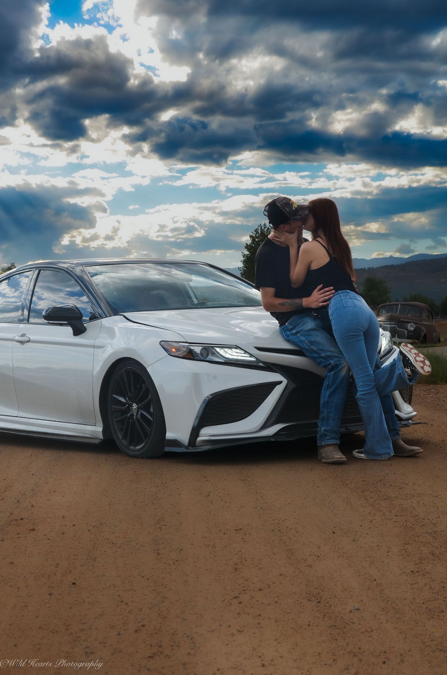 Creative outdoor portrait of a couple posing beside a white sports car beneath dramatic cloudy skies during an automotive photo session.