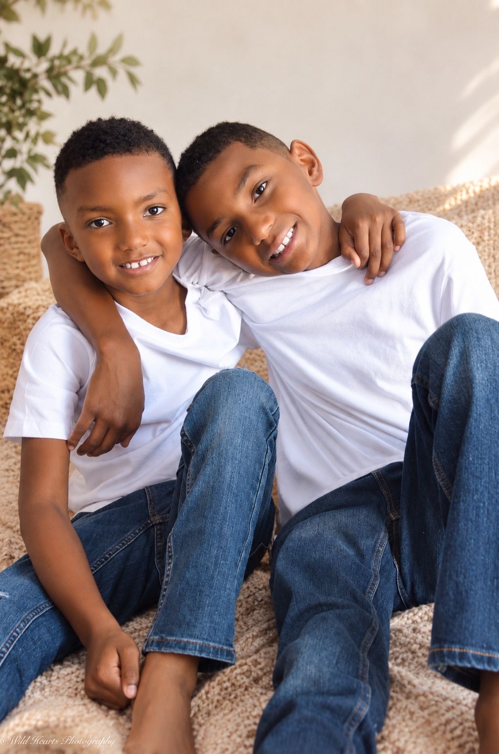 Two young brothers smiling together in matching white shirts and blue jeans during a sweet indoor sibling portrait session.