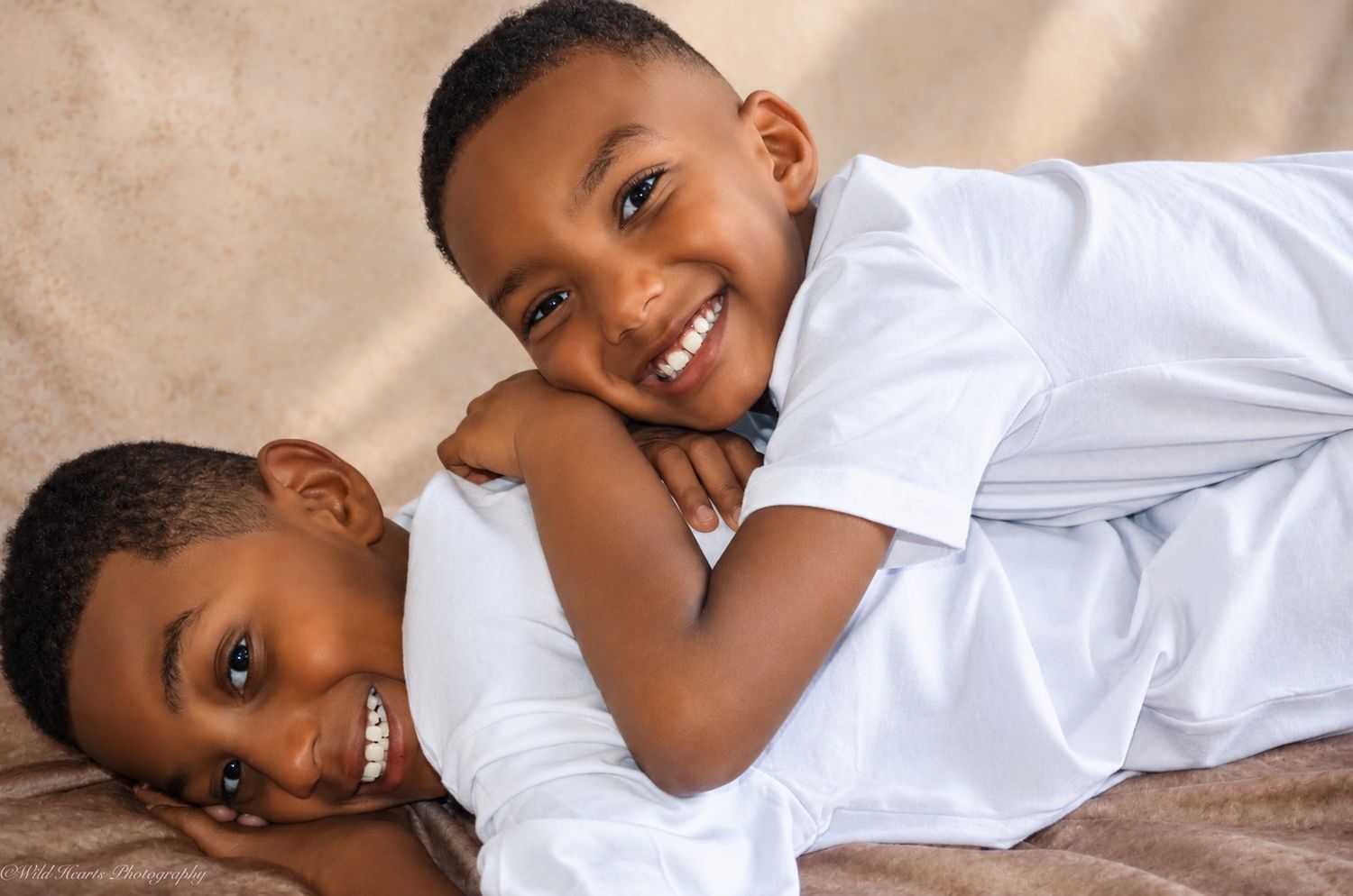 Two smiling young brothers wearing white shirts, lying together on a soft beige blanket during a children's portrait session.