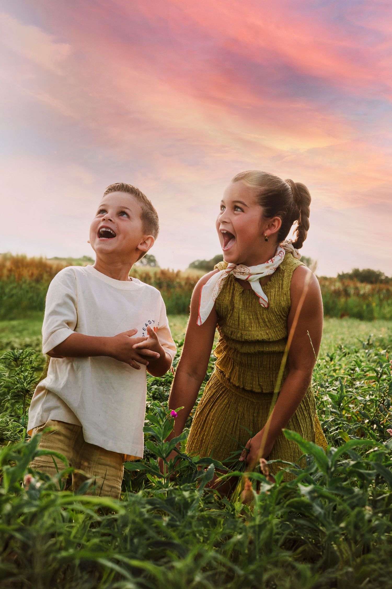 Two children laugh joyfully together while sitting in a lush green field during a vibrant sunset.