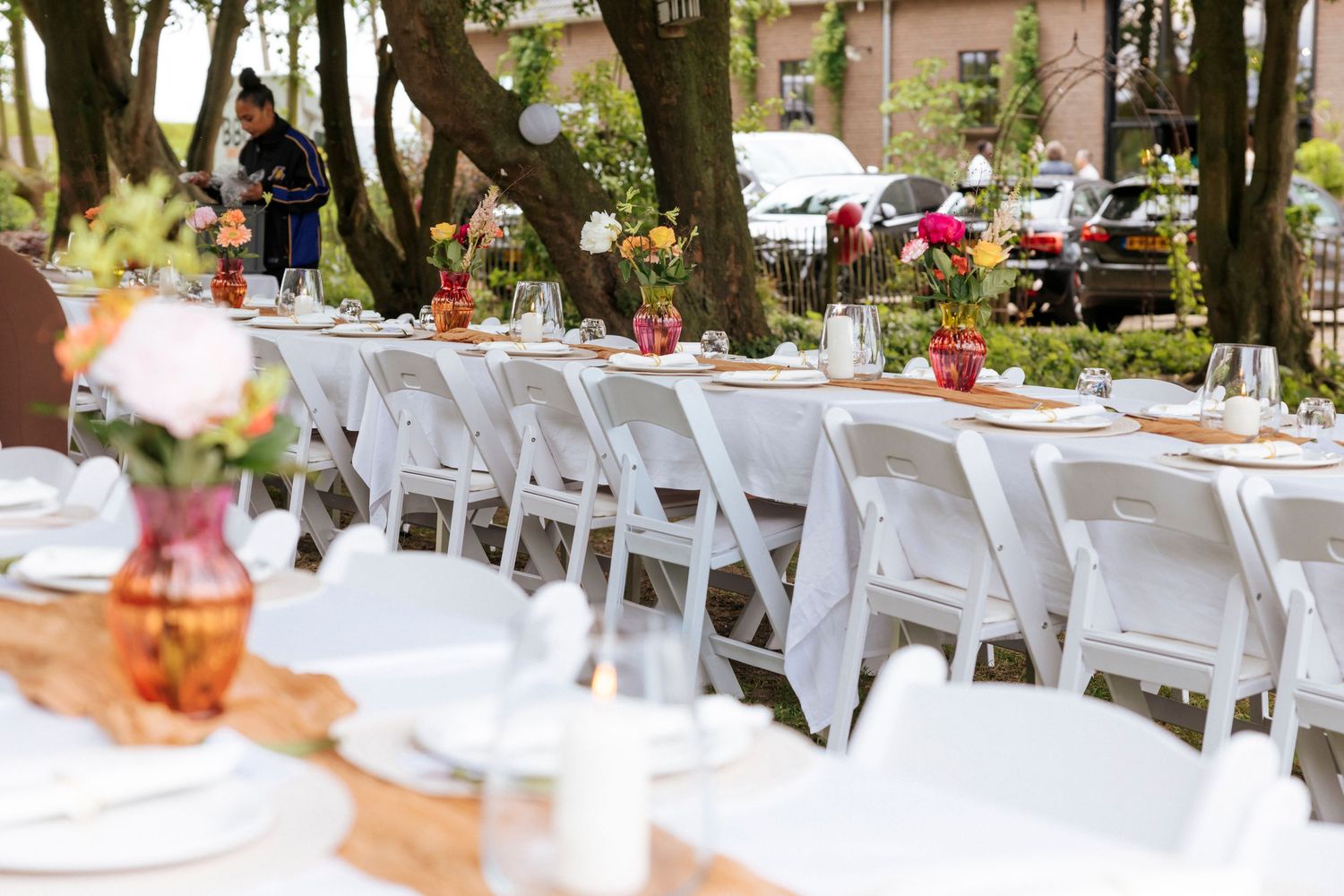 Long banquet tables with white folding chairs and floral centerpieces set up outdoors among trees for a garden event.