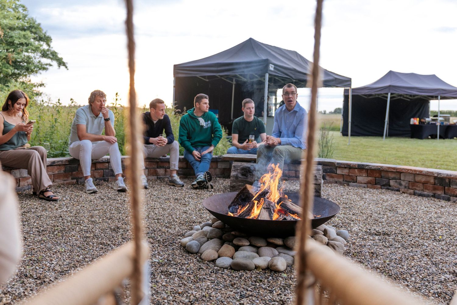 People gathered around a cozy campfire under pop-up canopies at an outdoor gravel patio area during dusk.