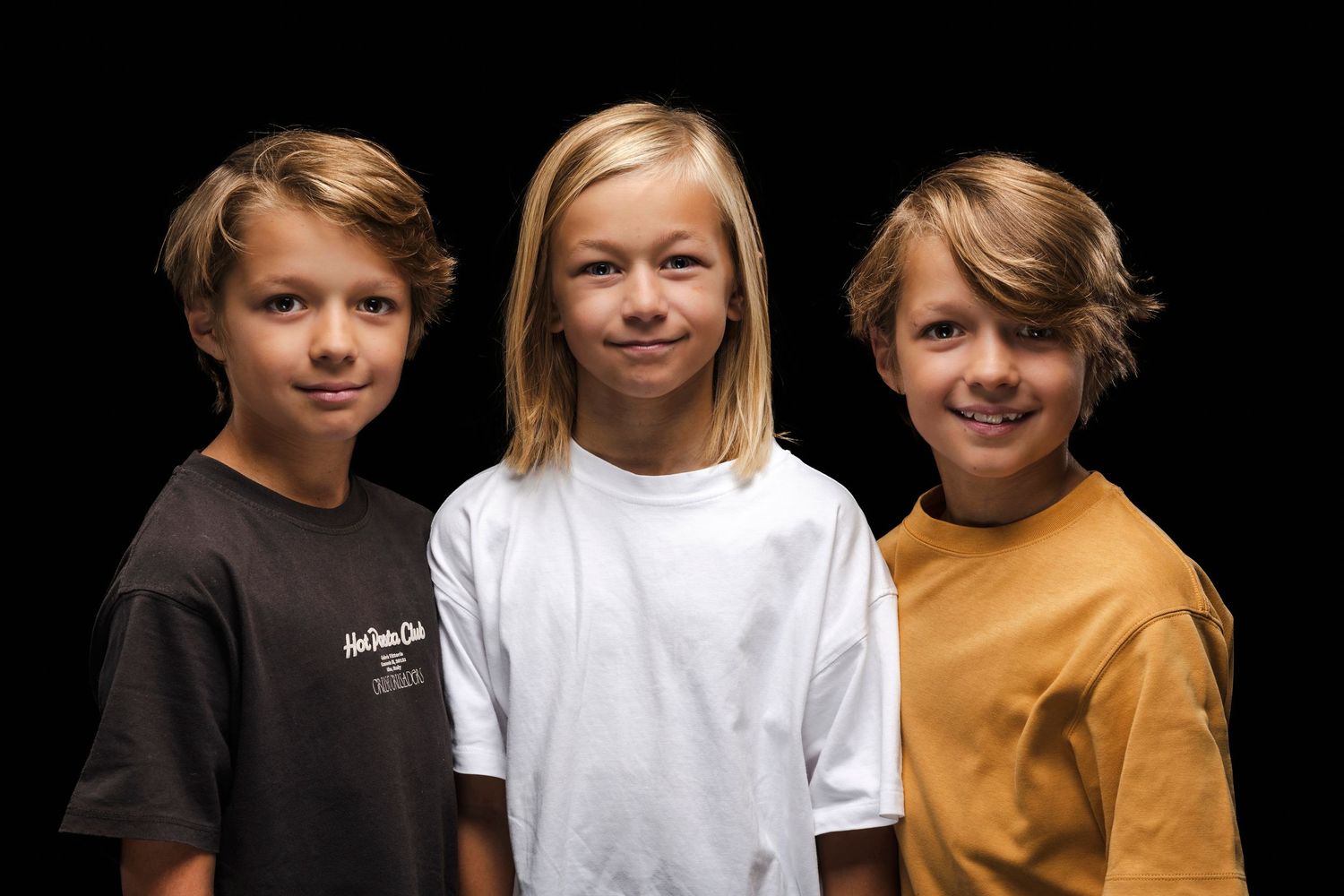 Studio portrait of three young people in white, brown and black t-shirts against dark background.