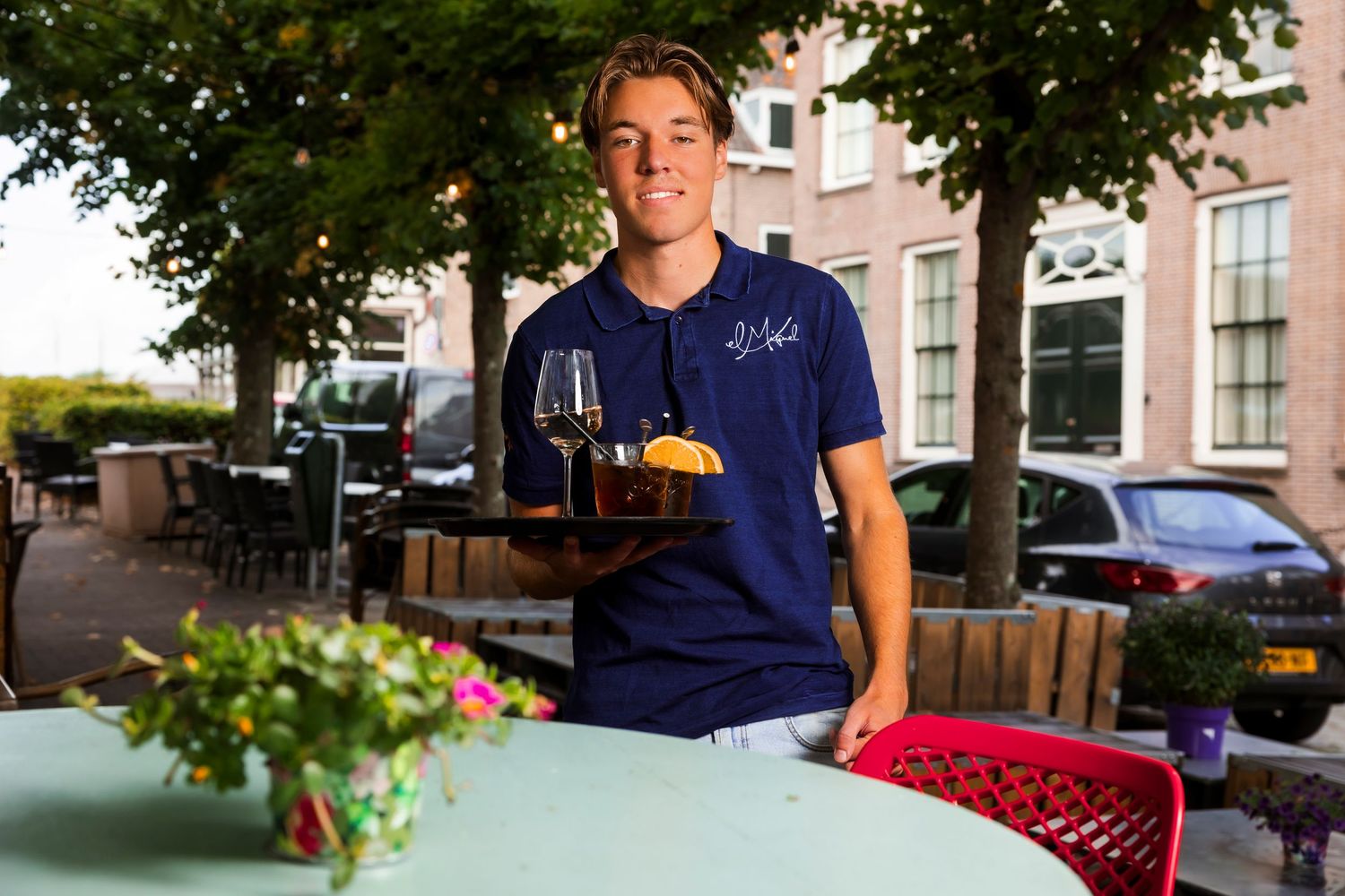 A server in a navy blue uniform holds a glass of wine and food on a patio outside a restaurant.
