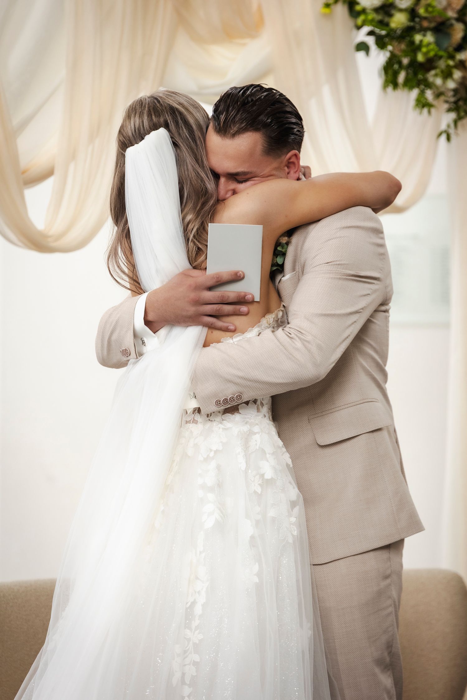 A romantic wedding moment captured as a couple embrace under elegant white draping and floral decor.