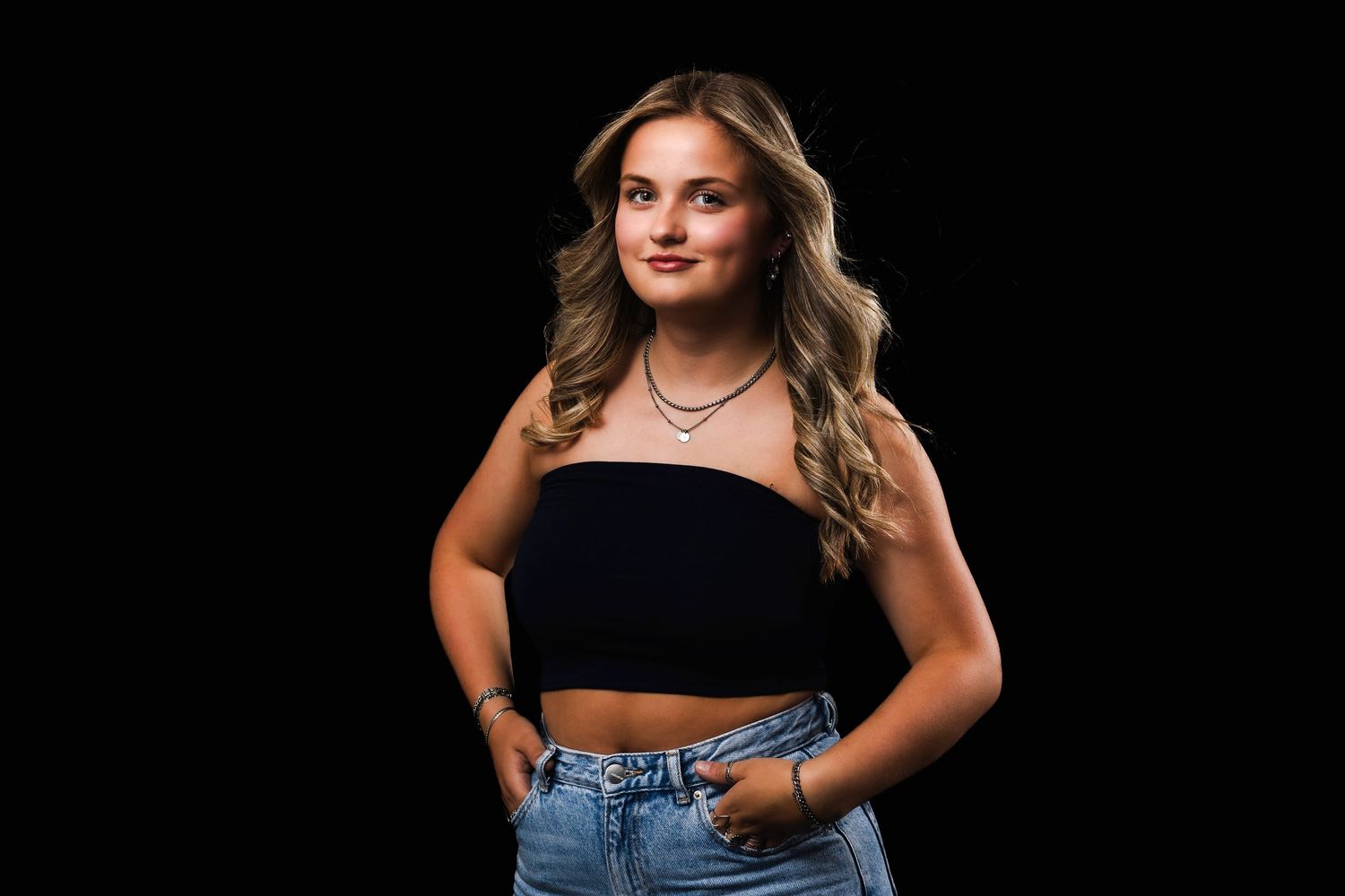 A figure poses confidently against a black background wearing a black tube top and high-waisted jeans.