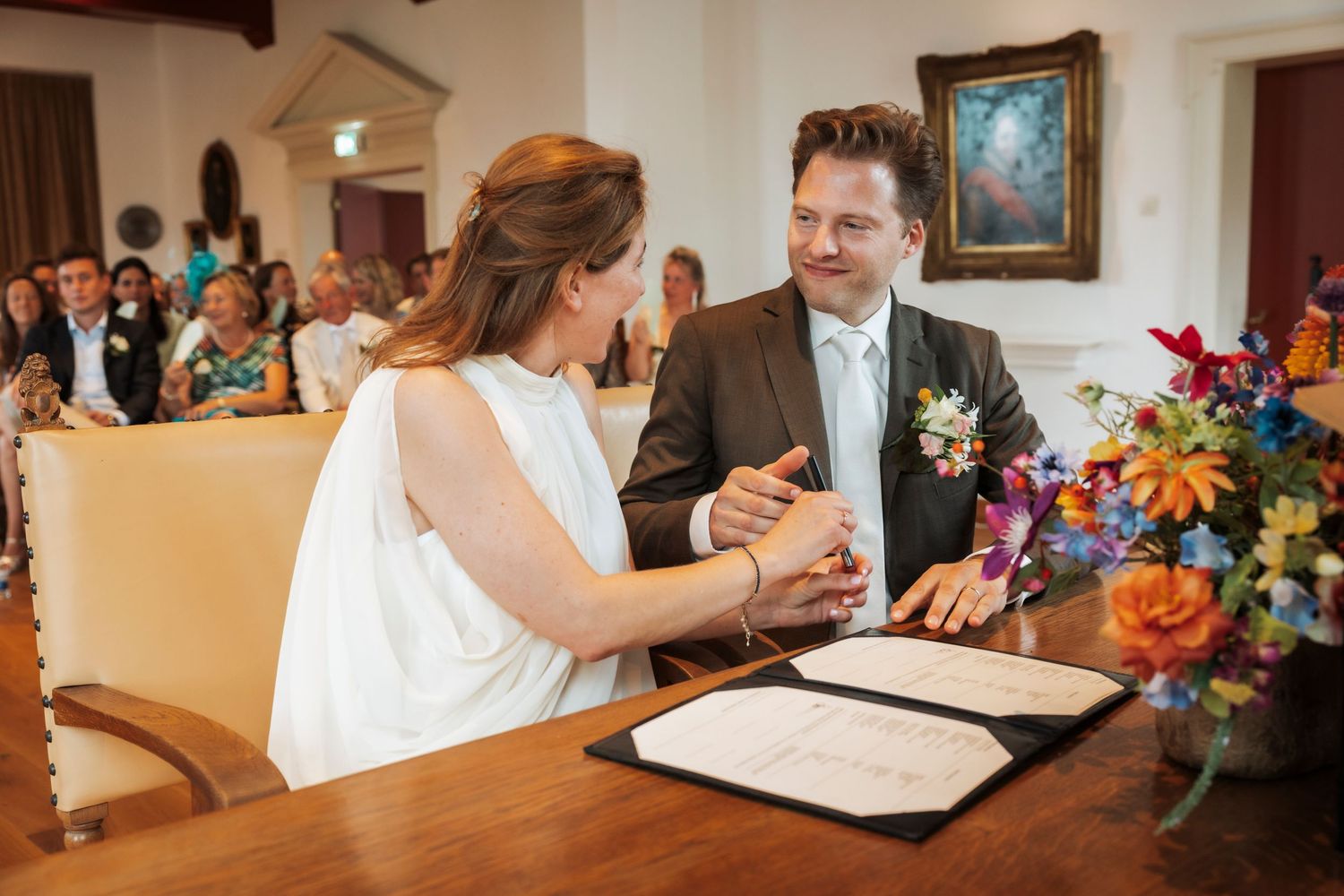 A couple signs their marriage certificate at a small ceremony with colorful floral arrangements in the background.