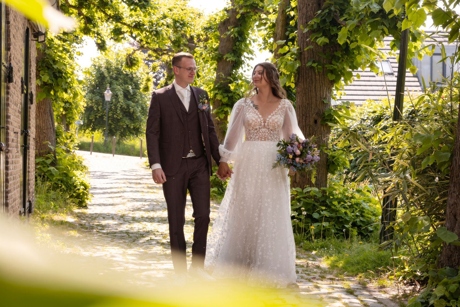 Newlyweds walking down a sunlit outdoor path surrounded by lush greenery and stone walls during a summer wedding.