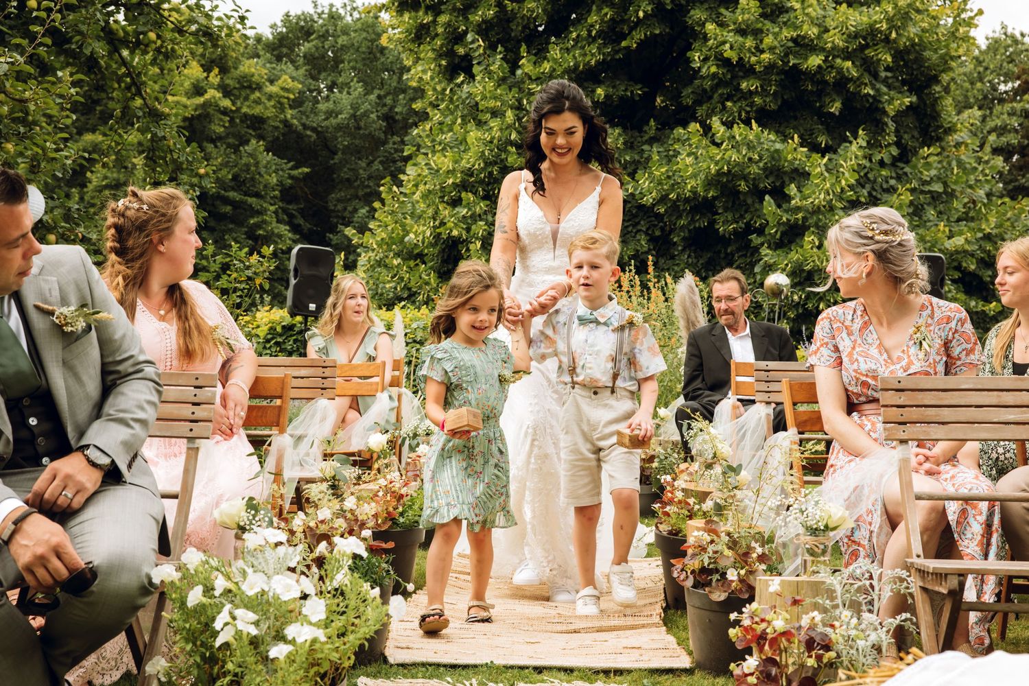 Wedding ceremony in an outdoor garden setting with wooden chairs and floral aisle arrangements in warm sunlight.