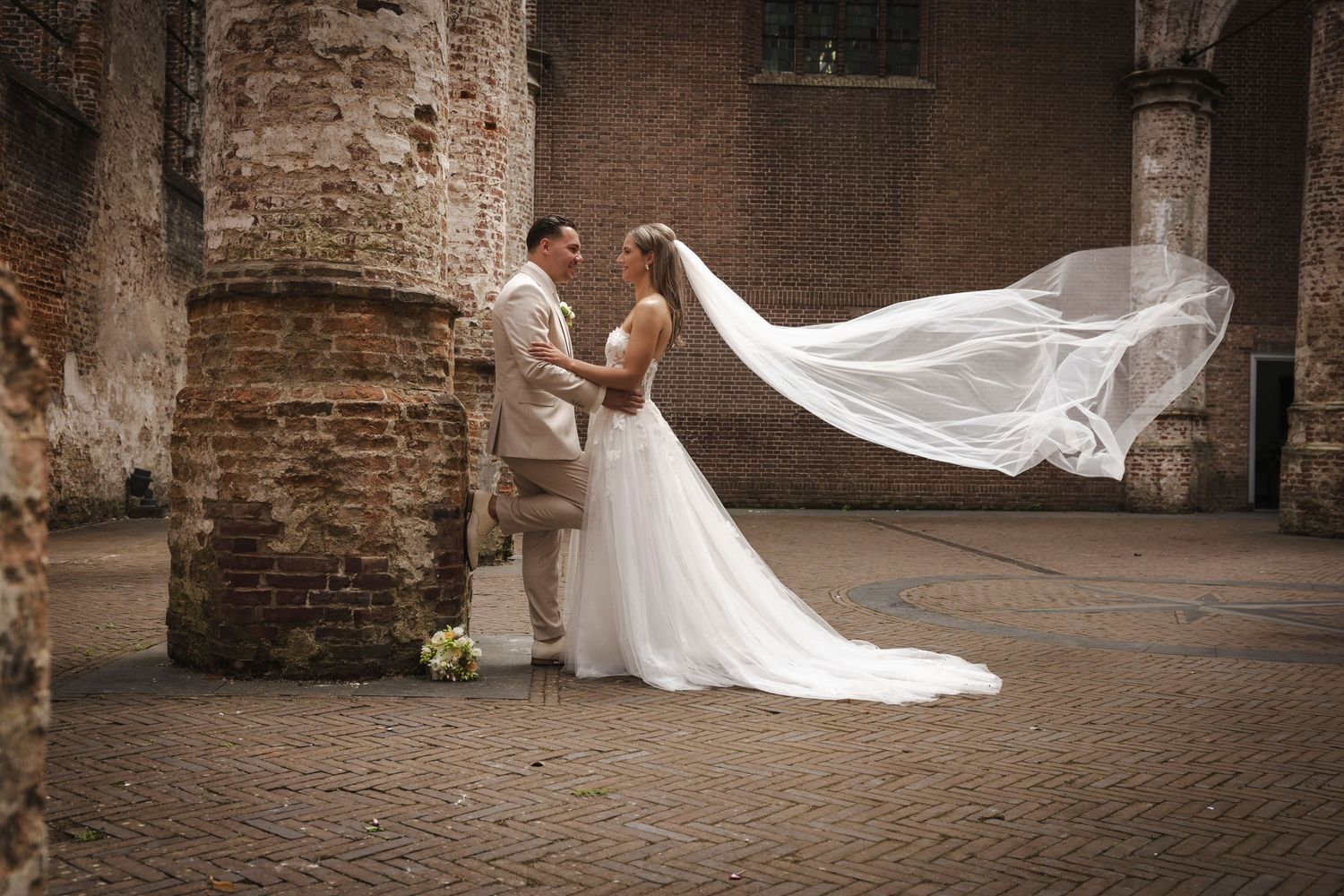 A romantic wedding photograph showing a veil flowing in the wind at a rustic brick venue with old stone columns.