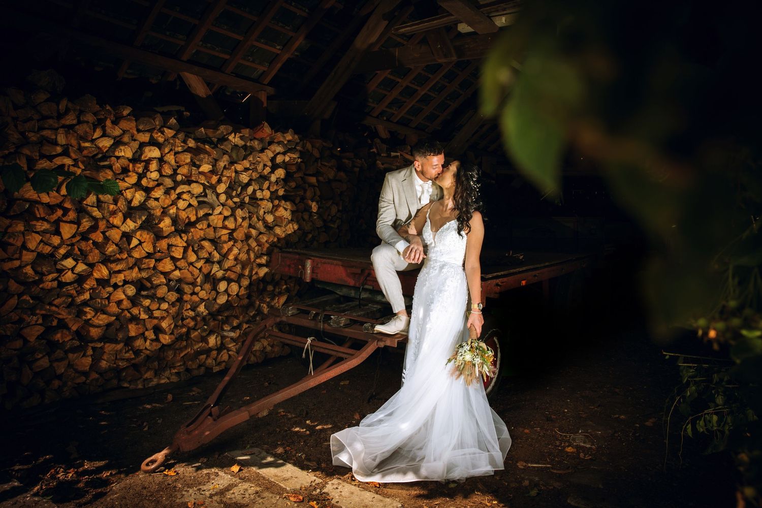 Couple in wedding attire pose intimately near stacked firewood at dusk in a rustic outdoor setting.