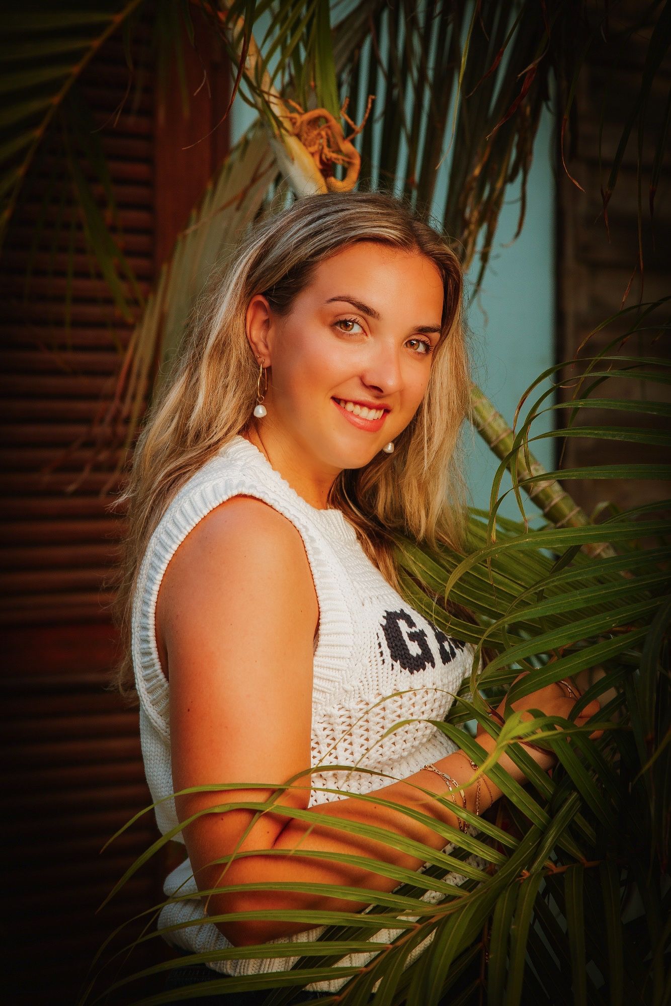A portrait in a tropical setting with palm fronds, featuring a white sleeveless top against a warm-toned background.