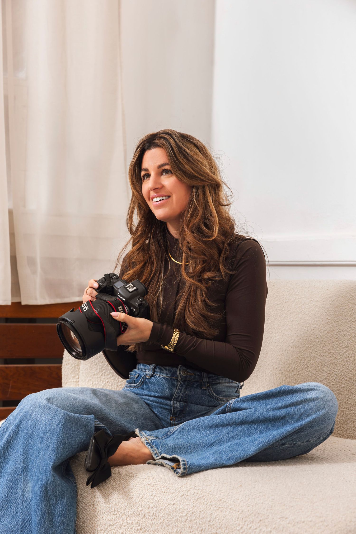 A person wearing jeans and a dark sweater sits casually on a light colored couch while holding a camera.