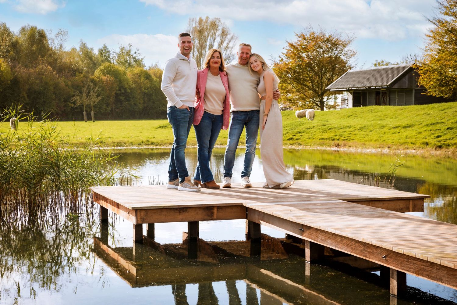 Three people standing together on a wooden dock overlooking a serene pond with autumn trees and a barn in the background.
