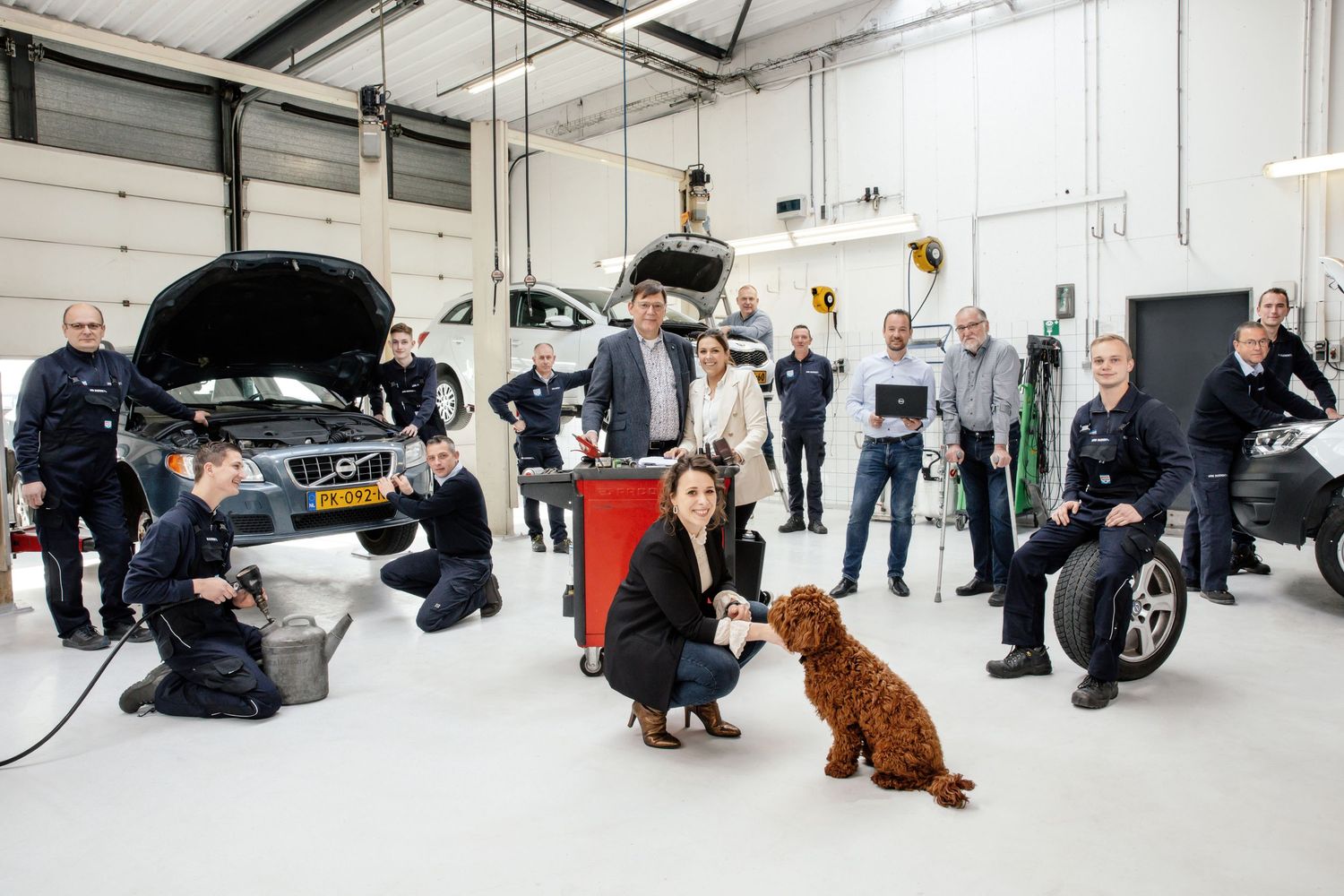 People gather in an automotive workshop with an open car hood and a brown dog in the center of the space.