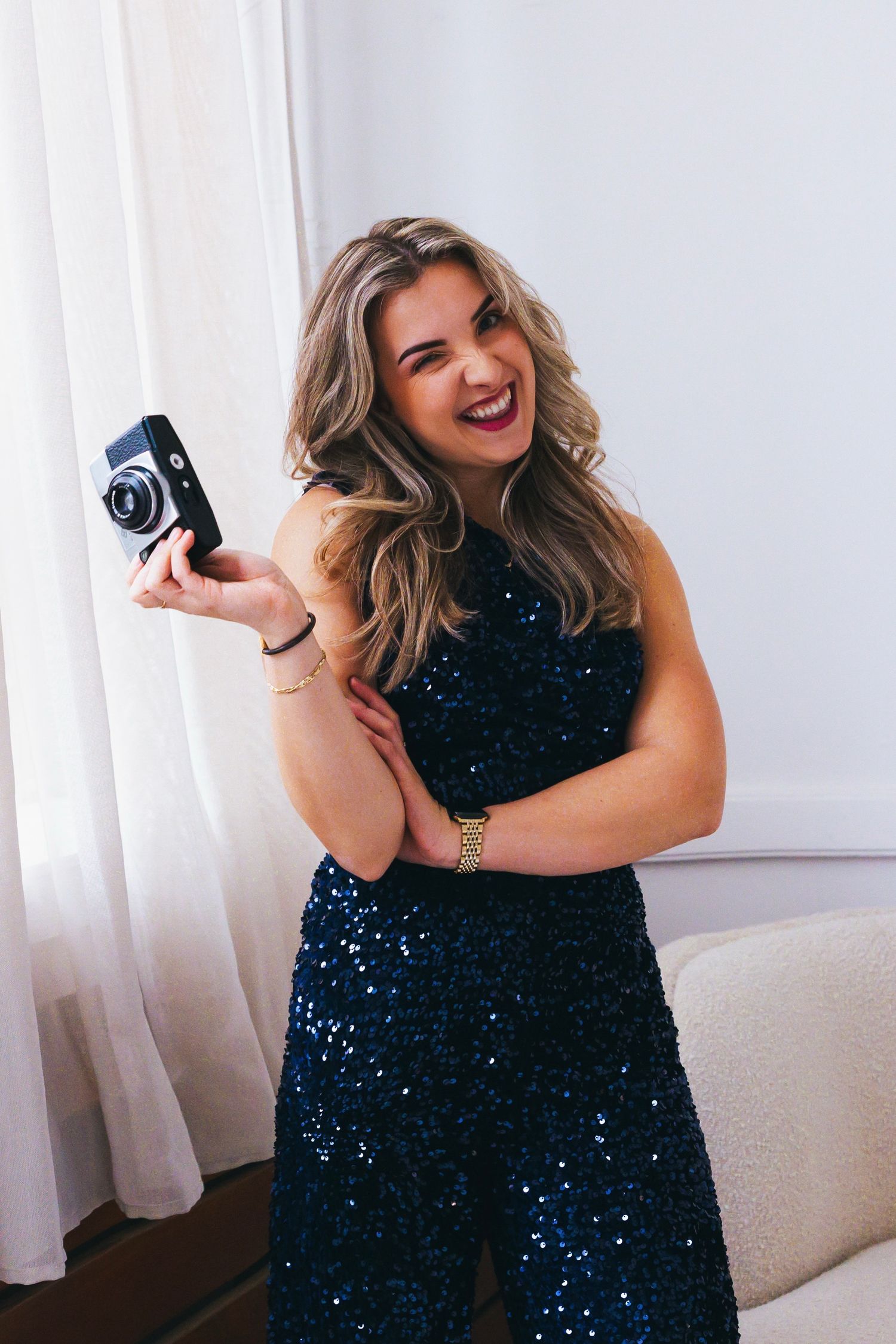 Photographer in sequined dress laughs while holding vintage camera in bright room.