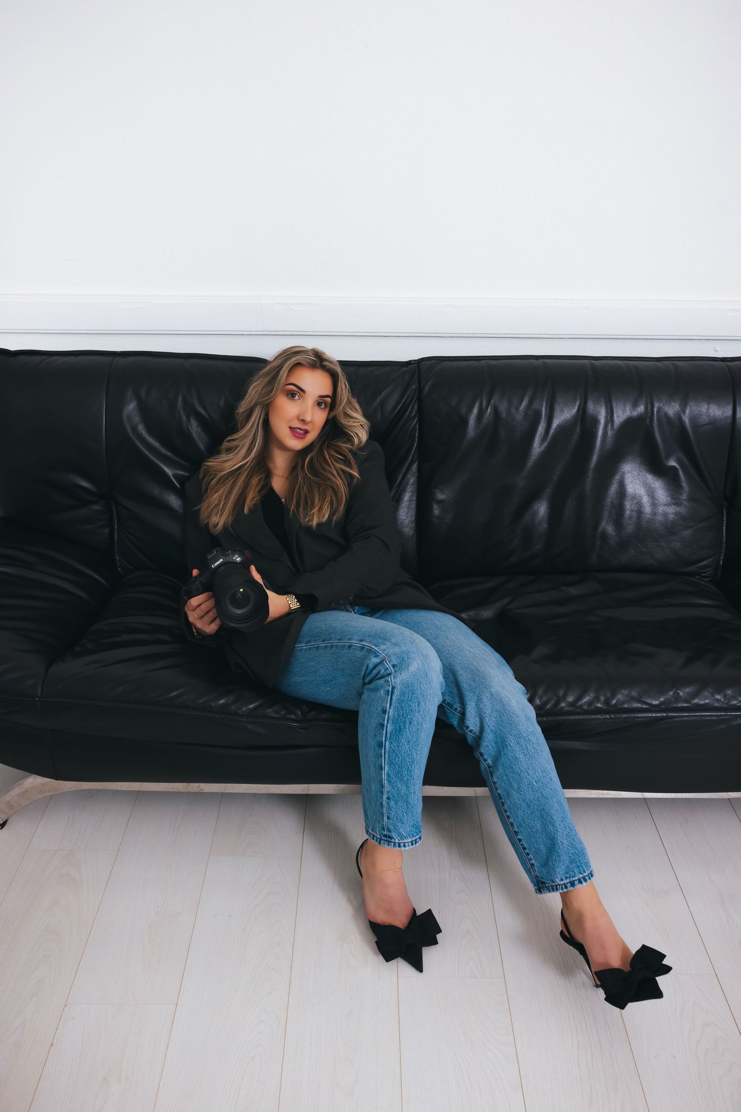 Photographer in casual attire holds camera while relaxing on black leather couch.
