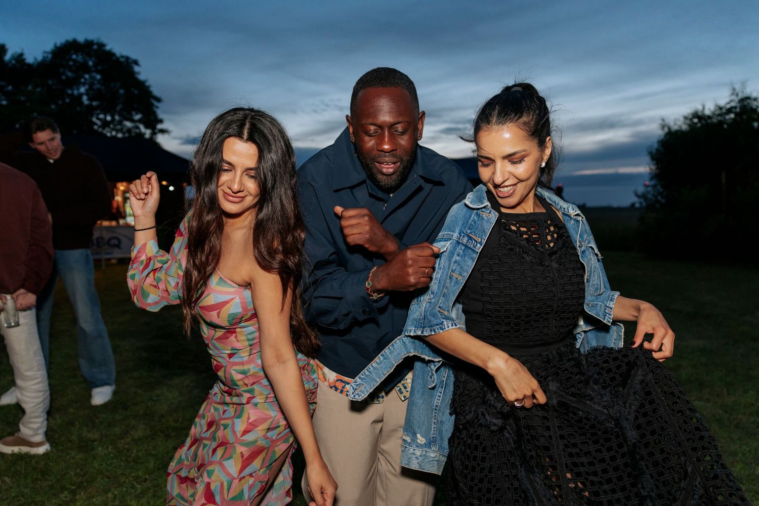Friends dancing together outdoors at a summer evening party, wearing casual dresses and denim jackets.