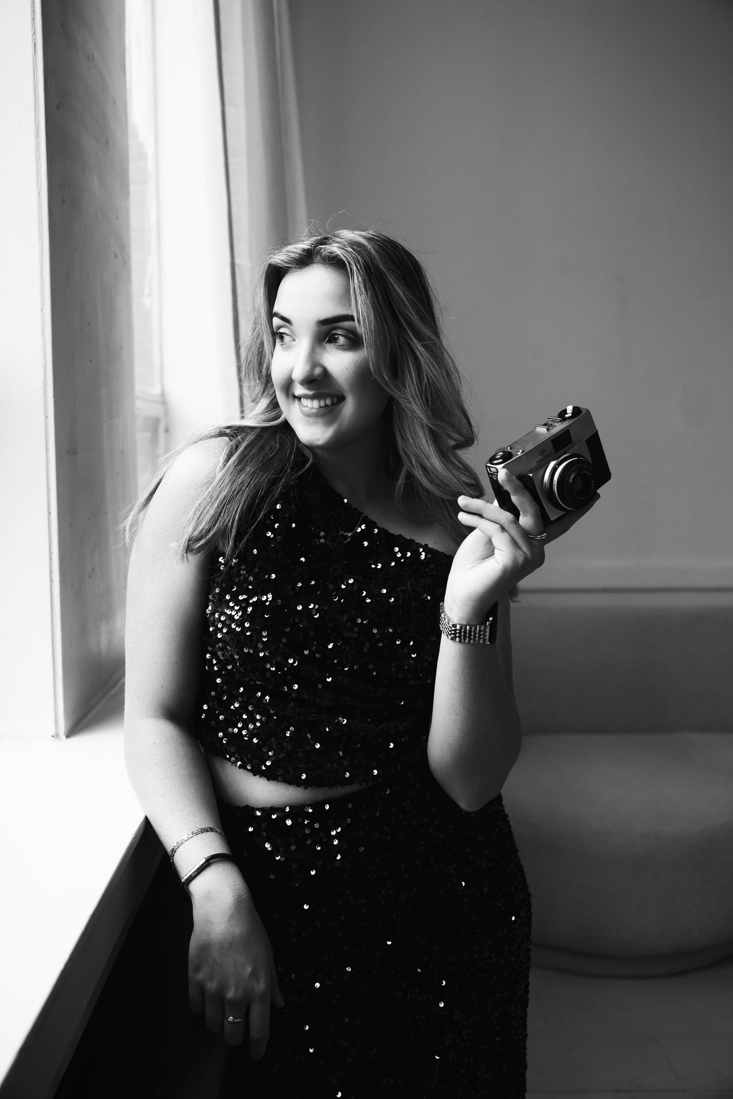 Professional photographer poses with camera near window in sparkly black formal dress.